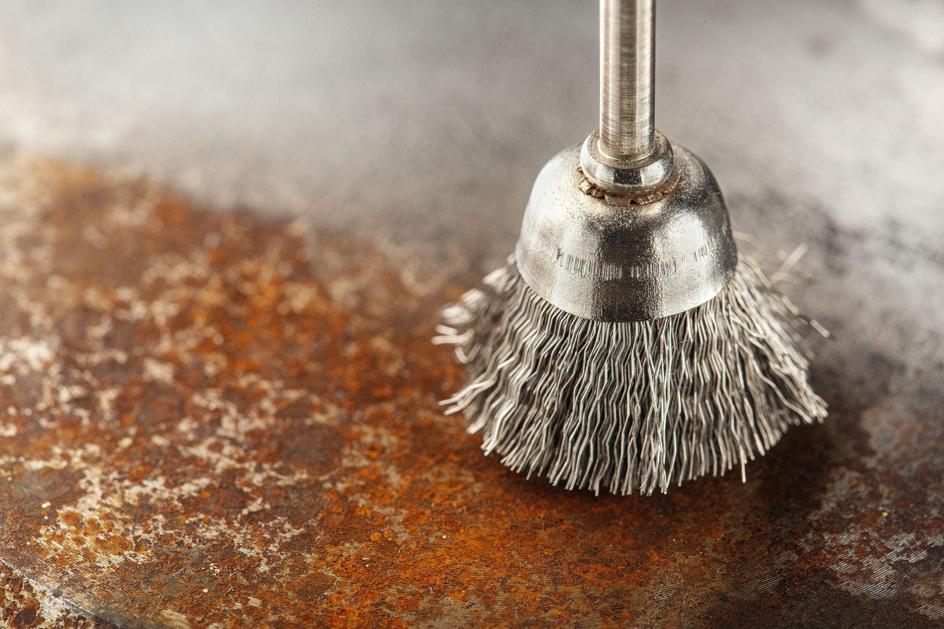 A wire brush removing rust from a brown, rusty metal surface.