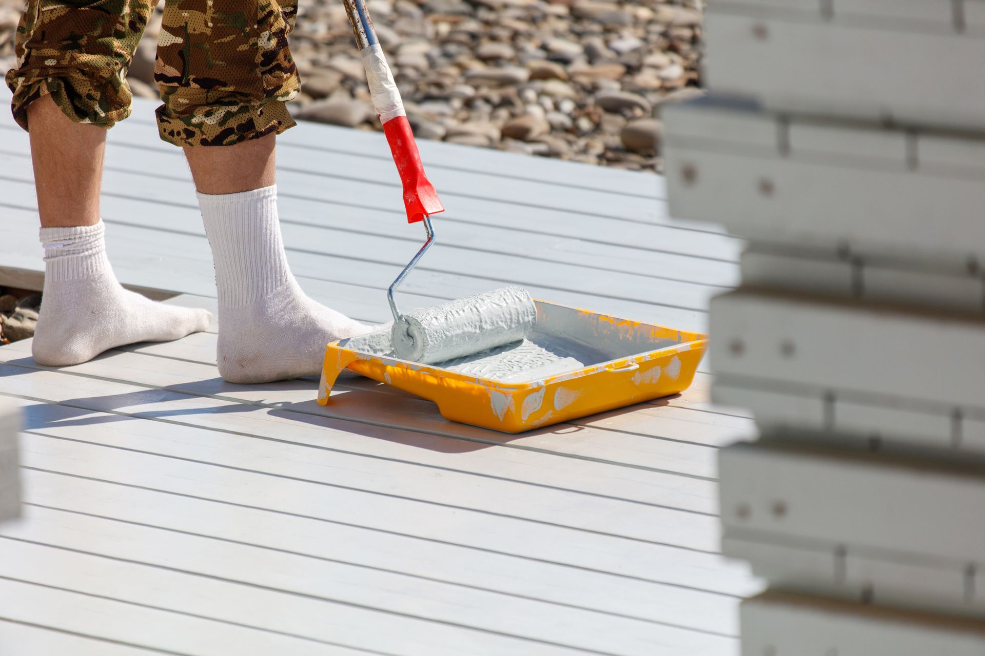 Person in camouflage pants and socks paints a deck with a roller and tray.