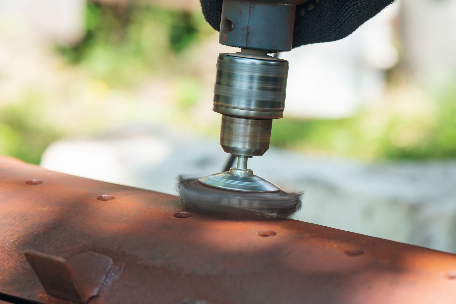 Person using a power drill with a wire brush to remove paint from rusty metal outdoors.