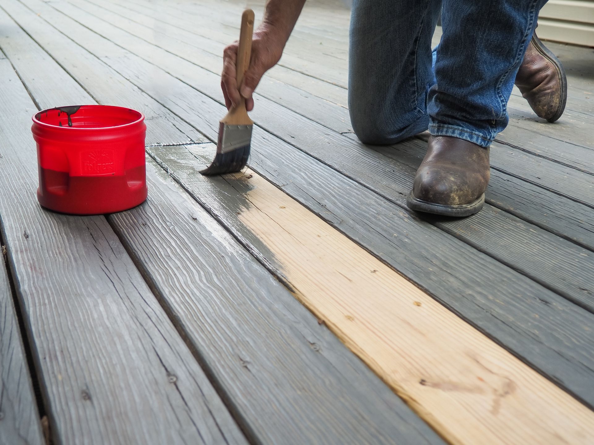 Person staining a wooden deck with a brush; red paint bucket nearby.