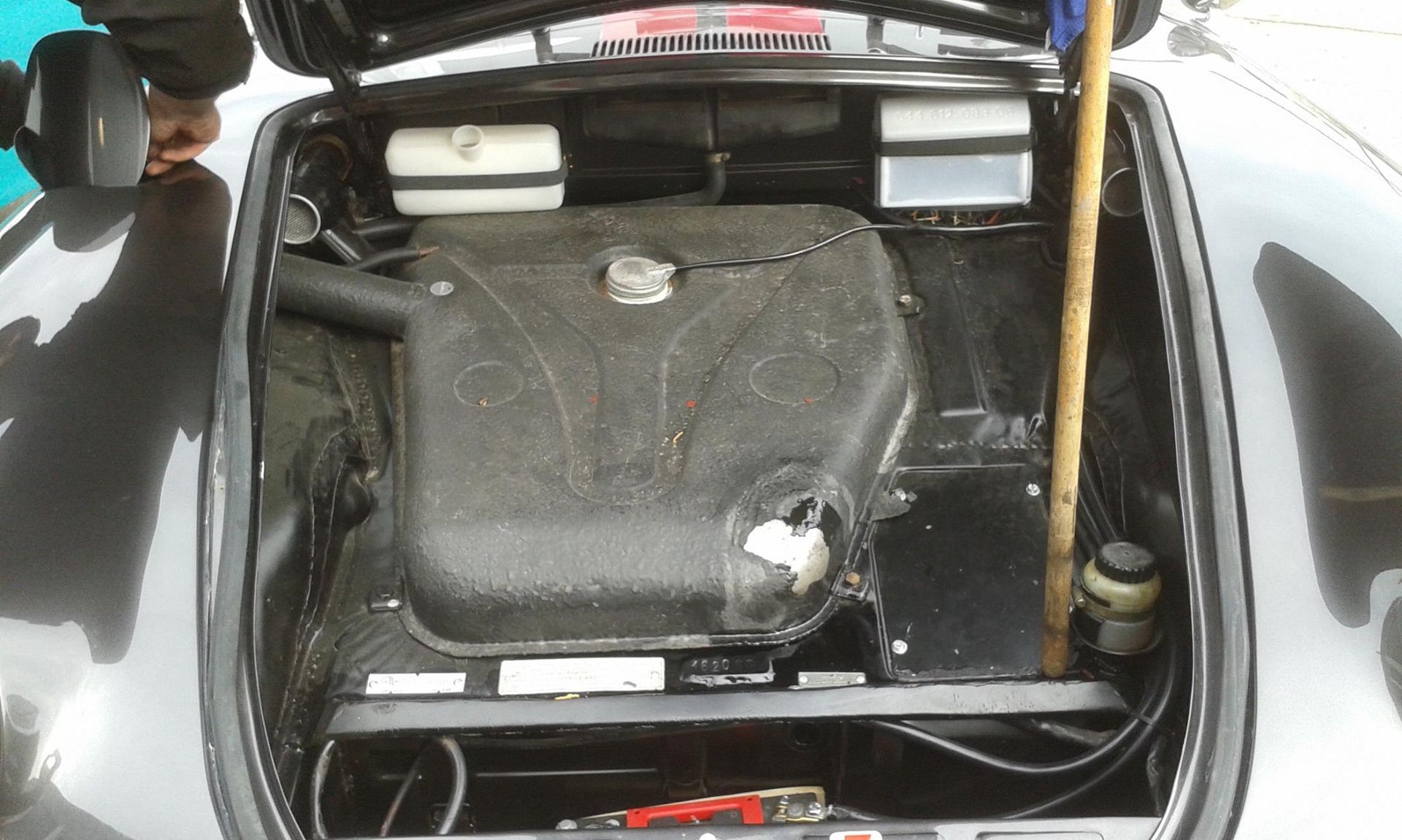 Black car engine compartment open; black, textured gas tank centered; two white reservoirs at top.