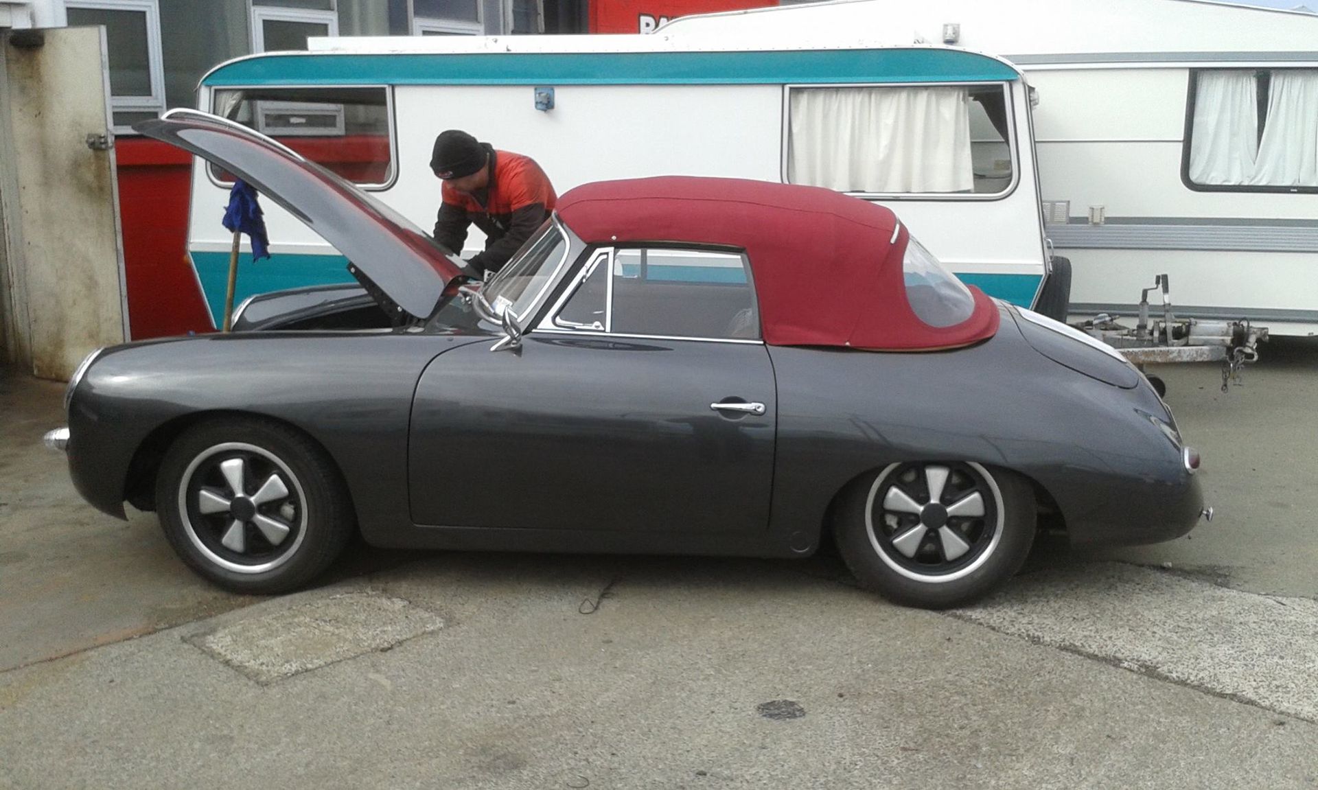 Dark gray convertible with red top, hood open, person working on engine, parked next to a camper.