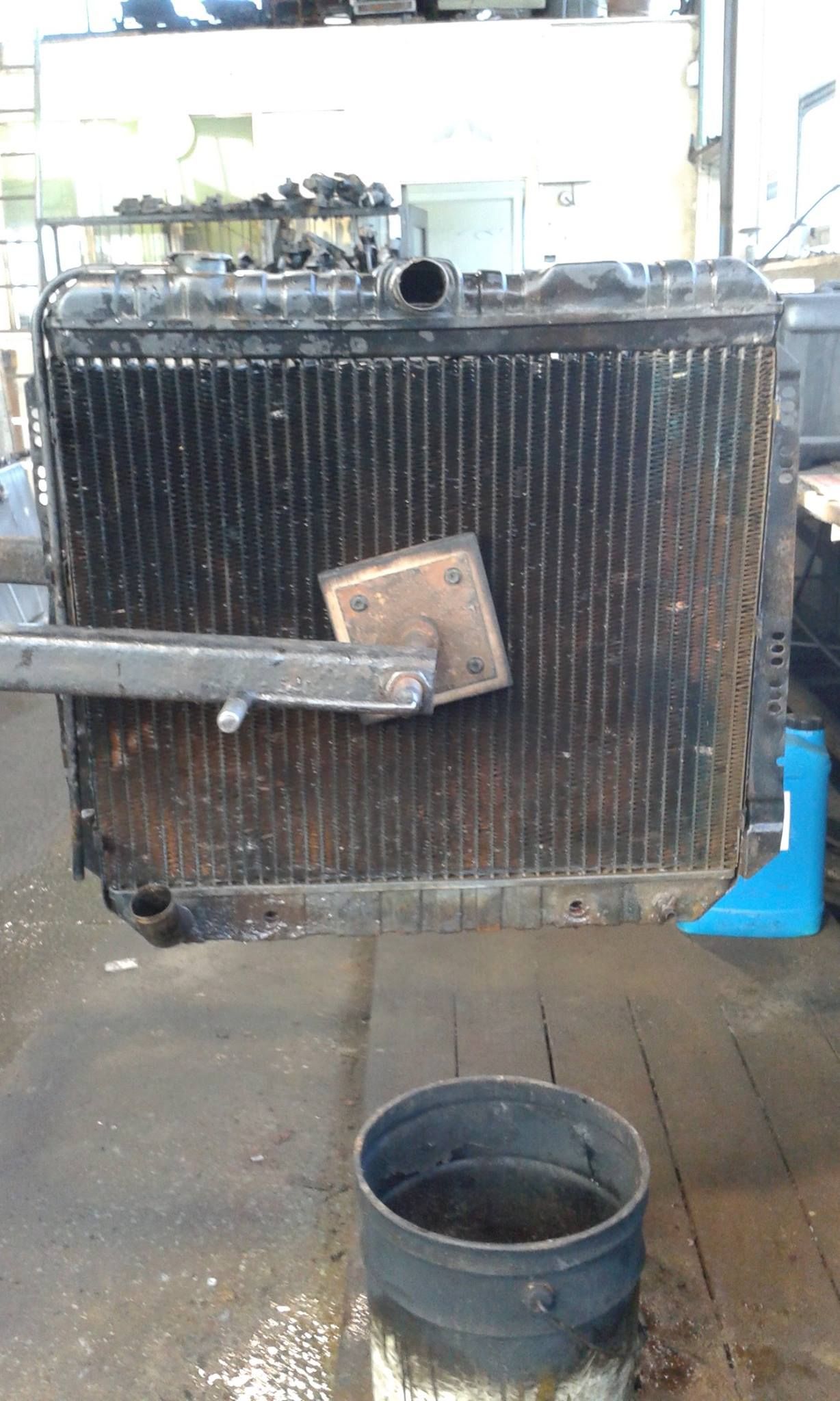 A mechanic working on a rusty car radiator with a tool in a shop setting.