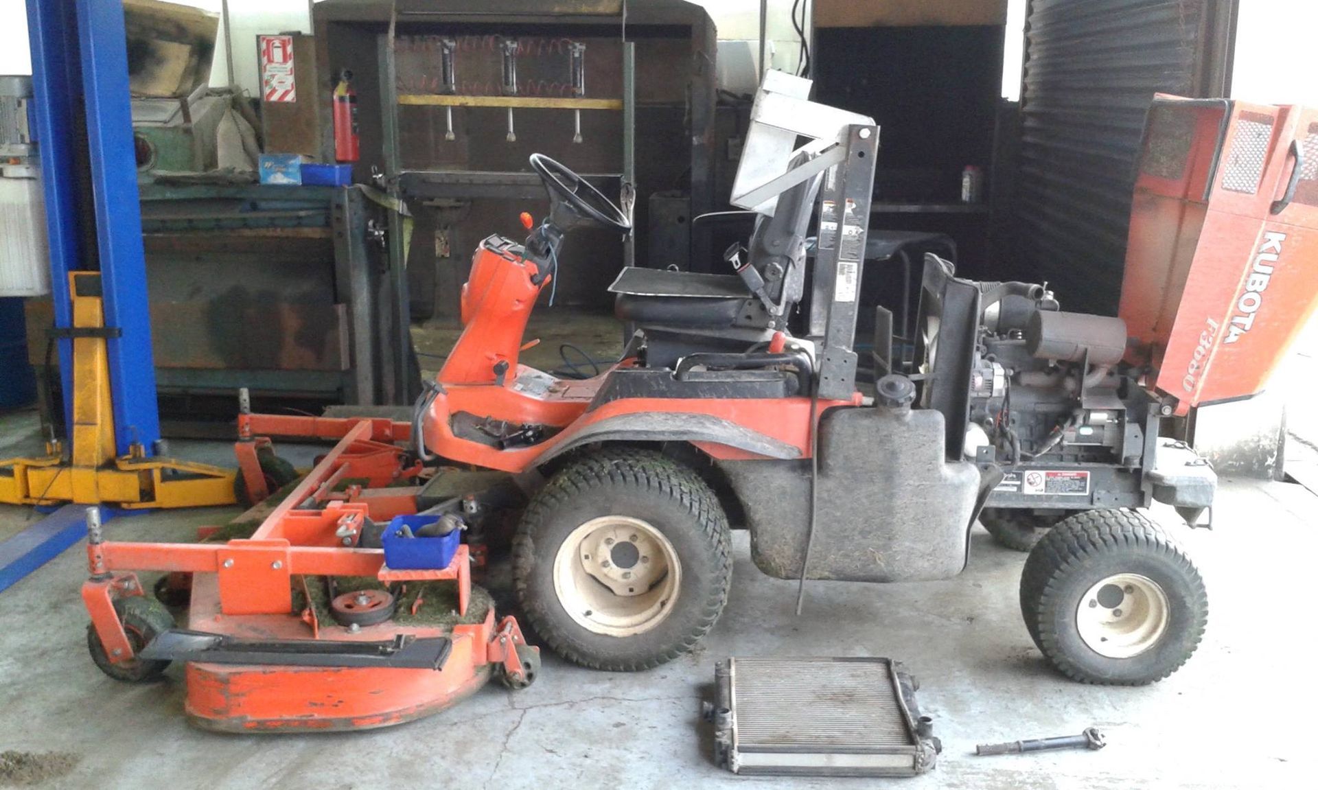 Orange riding lawn mower in a garage, engine exposed, with a detached radiator and mower deck.