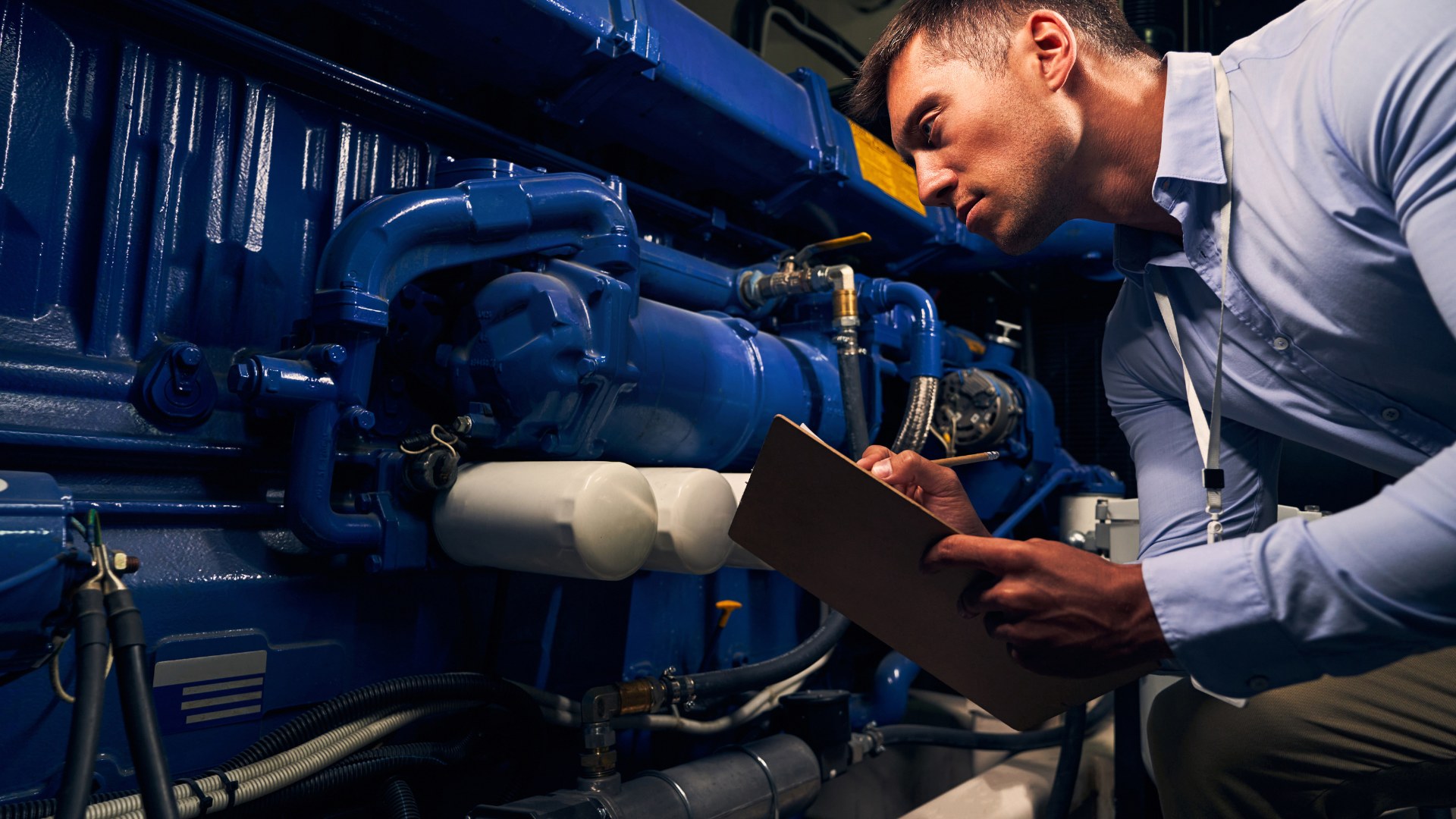 A man is kneeling down in front of a blue engine and writing on a clipboard.