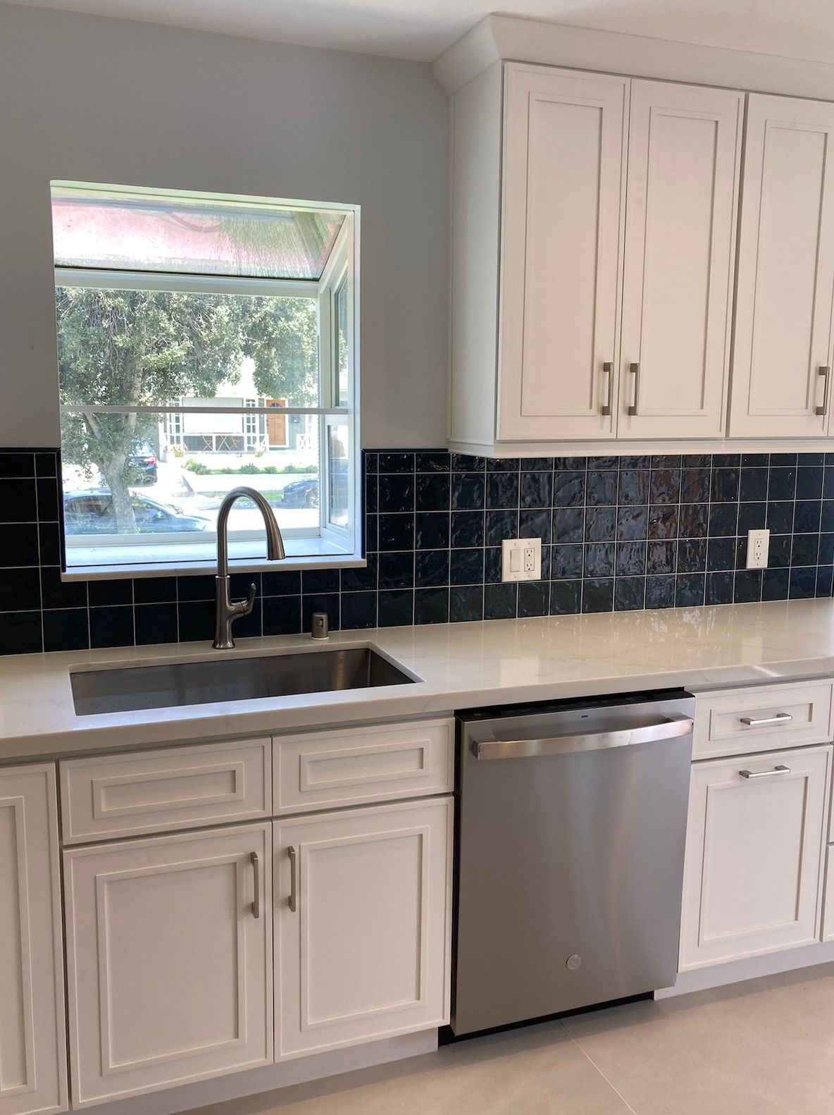 White kitchen cabinets, stainless steel dishwasher and sink, navy tile backsplash, and a window.
