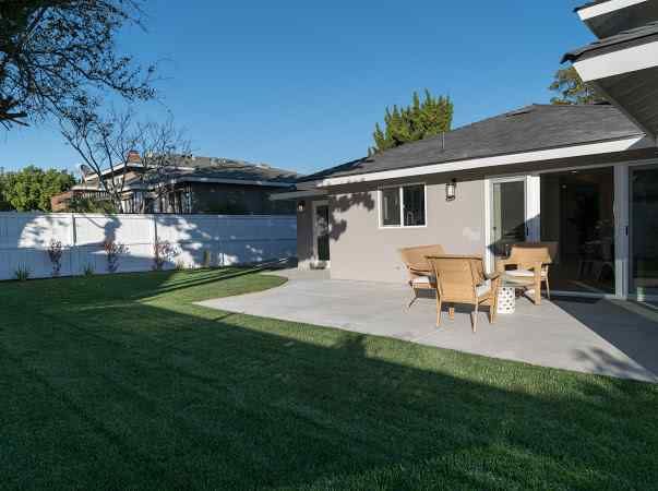 Backyard with patio, lawn, seating area, and white fence under a blue sky.