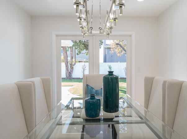 Dining room with glass table, white chairs, teal vases, and a chandelier; a view of the backyard through the windows.