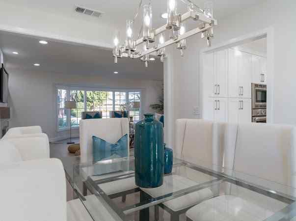 Dining room with glass table, white chairs, blue vase, and chandelier; kitchen in the background.