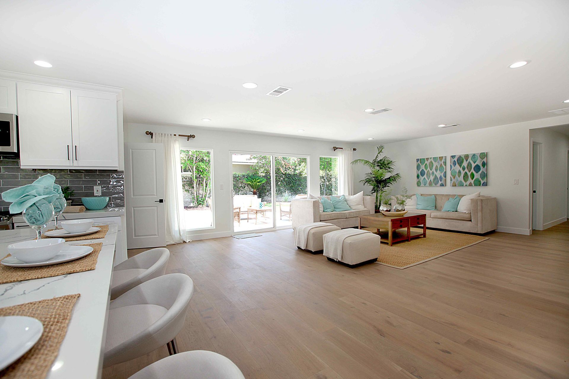 Bright open living space with kitchen and patio access; light wood floors, white walls, and neutral furniture.