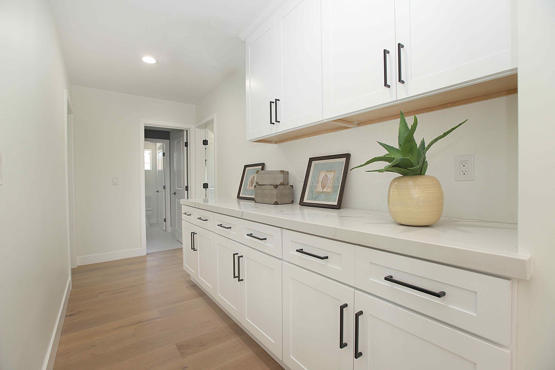 A bright white hallway with long white cabinets, black hardware, and decor. Light wood flooring.