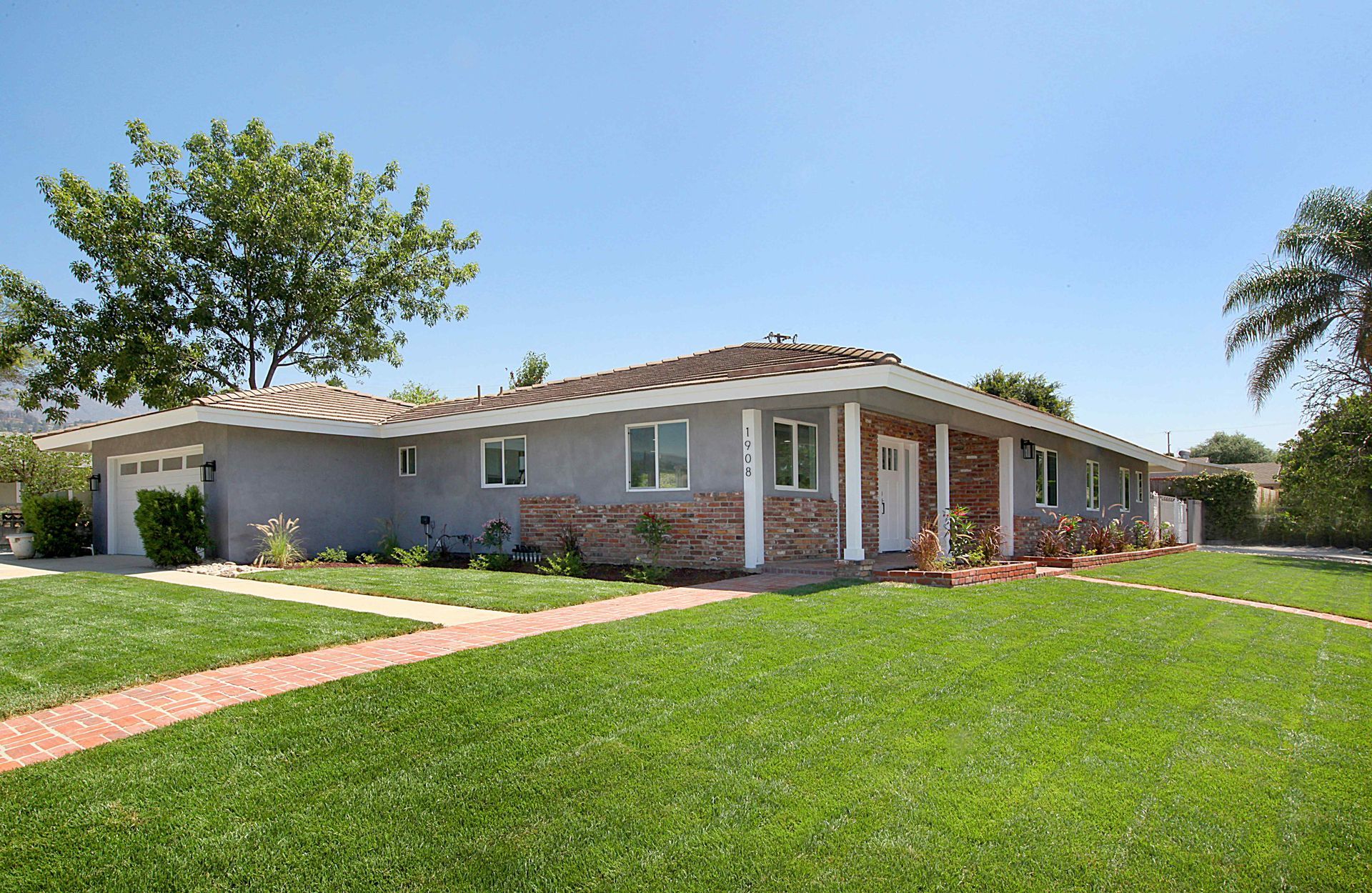 Ranch-style house with gray stucco and a brick walkway on a sunny day.