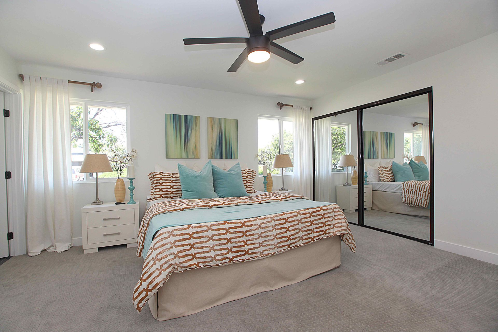 Bedroom with a bed, two windows, a large mirror, and a ceiling fan; light colors and teal accents.