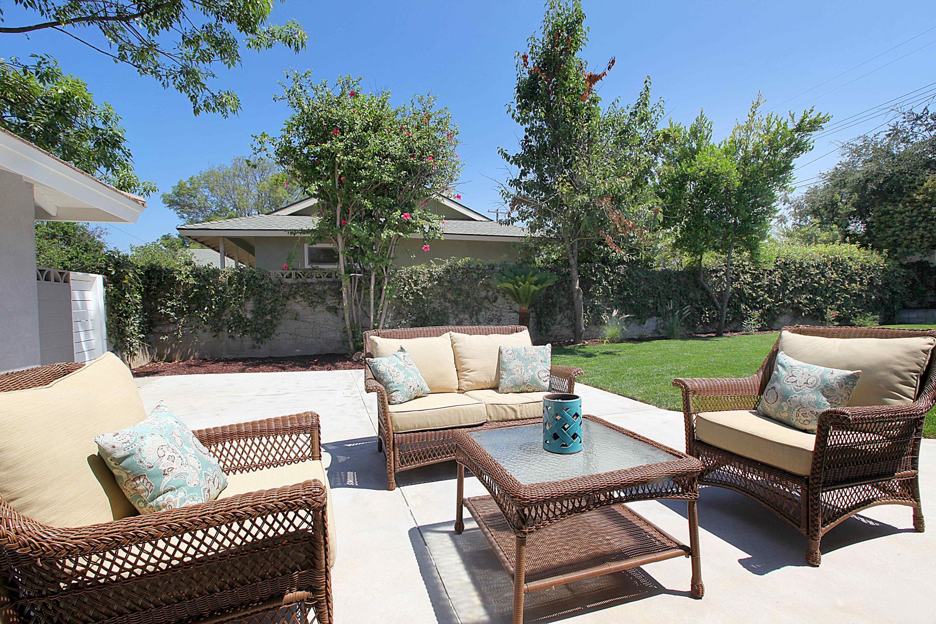 Patio with wicker furniture, tan cushions, and blue pillows in a grassy backyard.