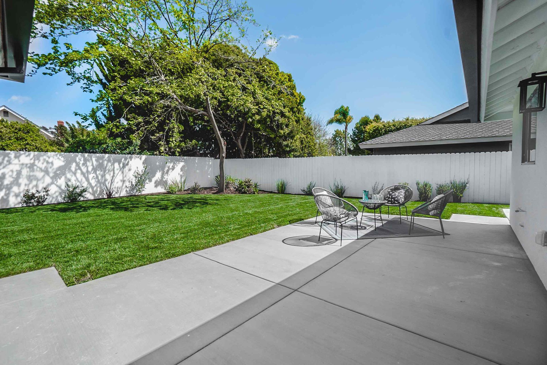Backyard with green grass, patio furniture on a gray patio, and white fence.