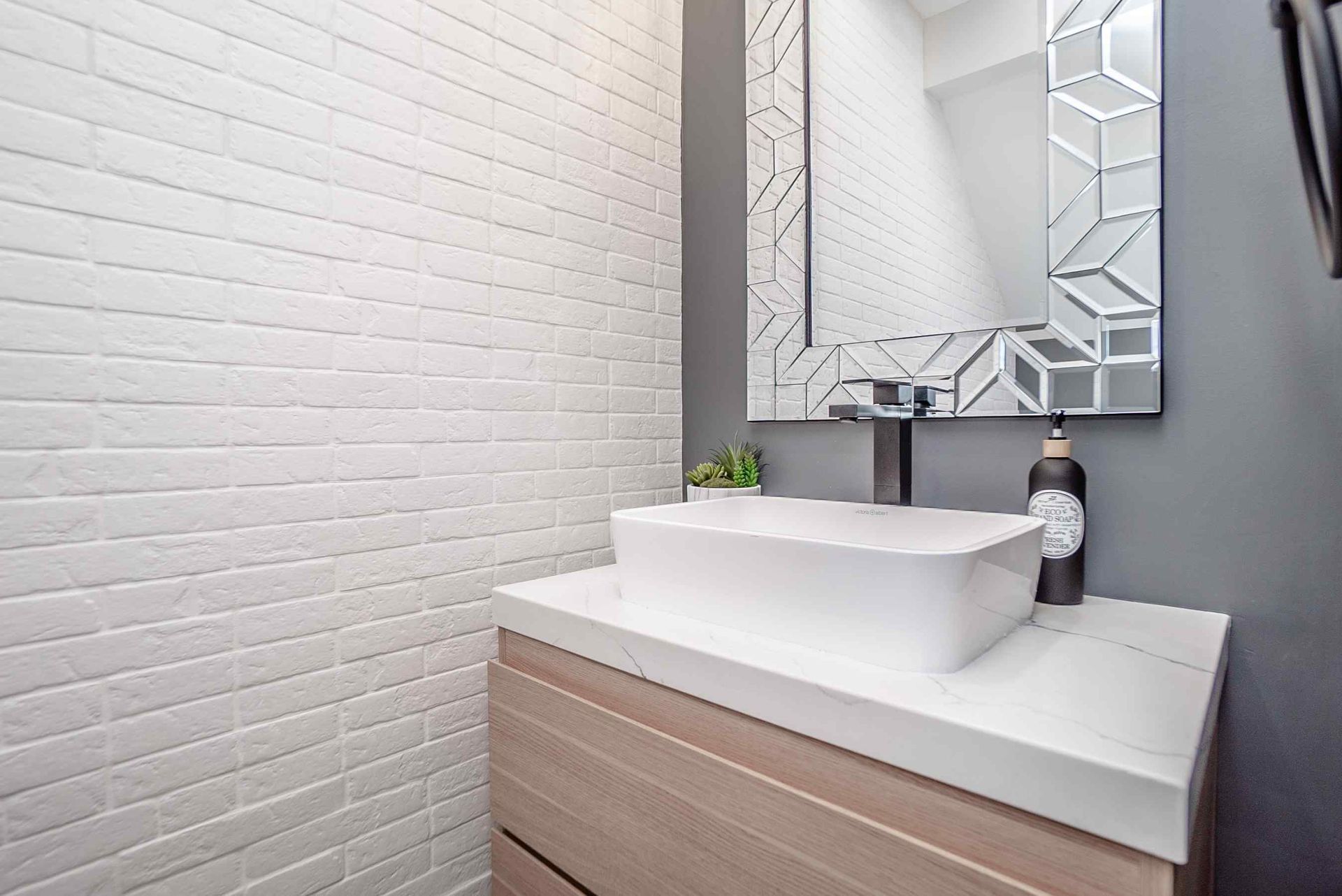 Modern bathroom with white sink, wooden cabinet, geometric mirror, and white brick wall.
