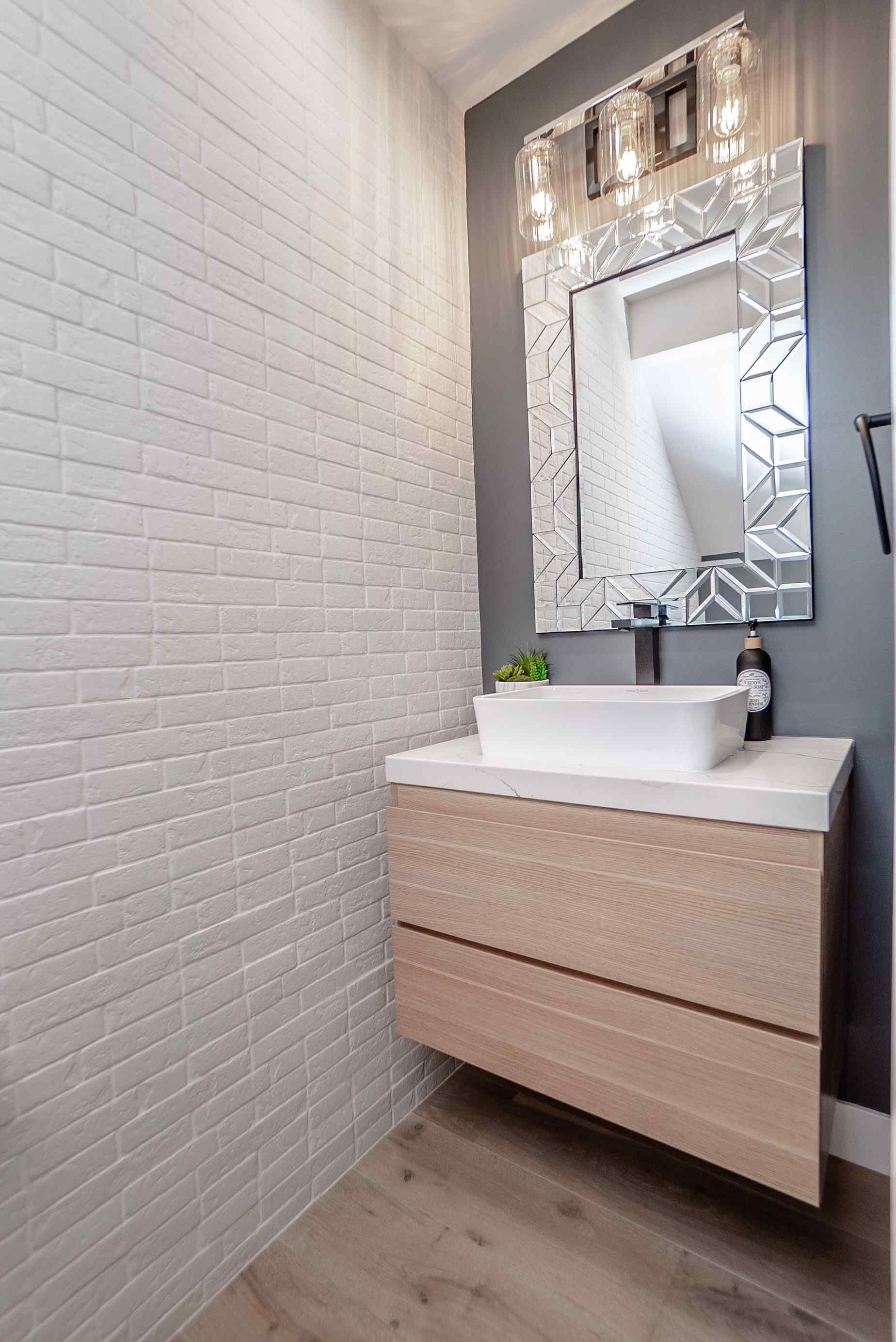 Modern bathroom with a floating wooden vanity, white sink, and patterned mirror. White brick tile wall.