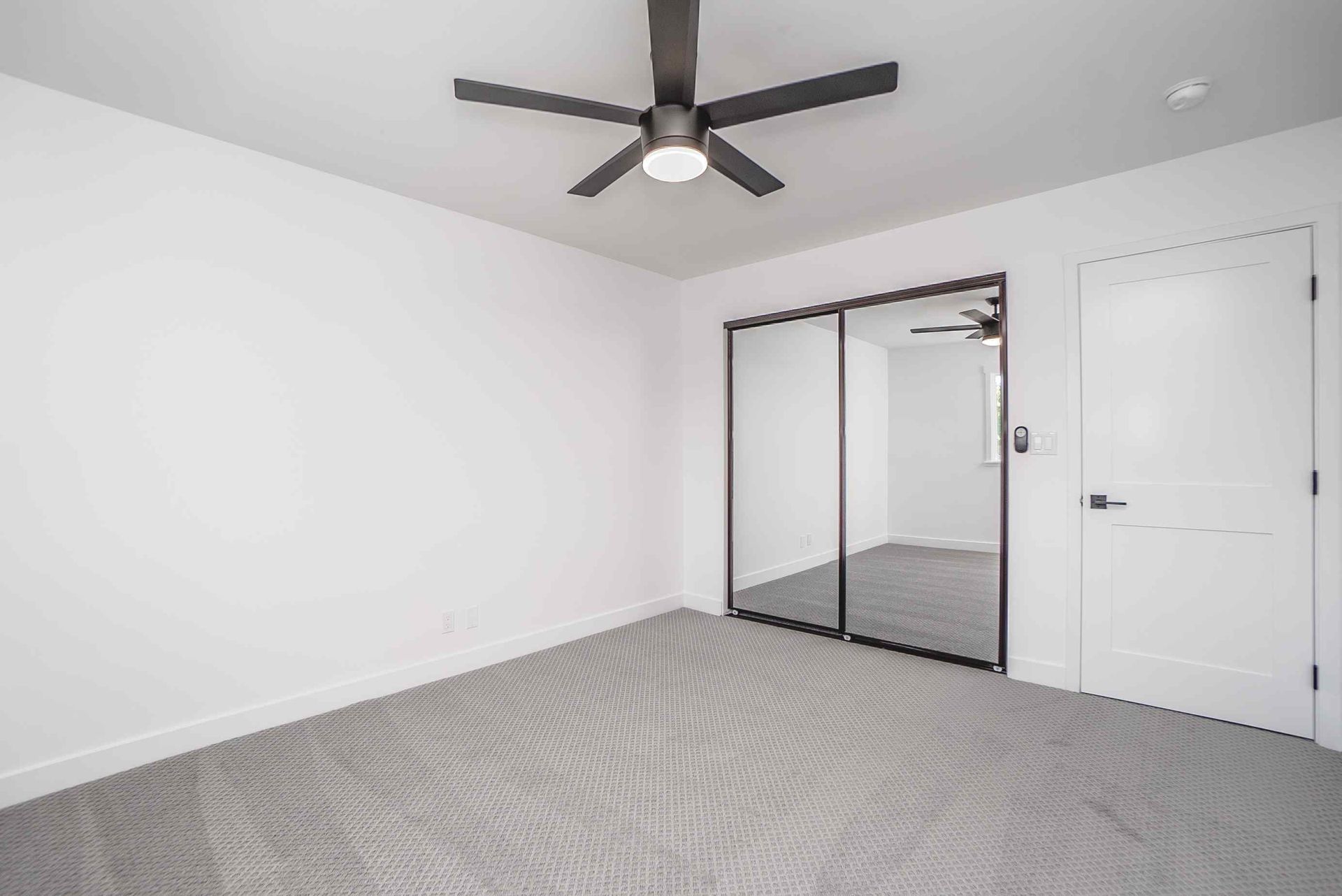 Empty bedroom with gray carpet, white walls, sliding mirrored closet doors, and a ceiling fan.