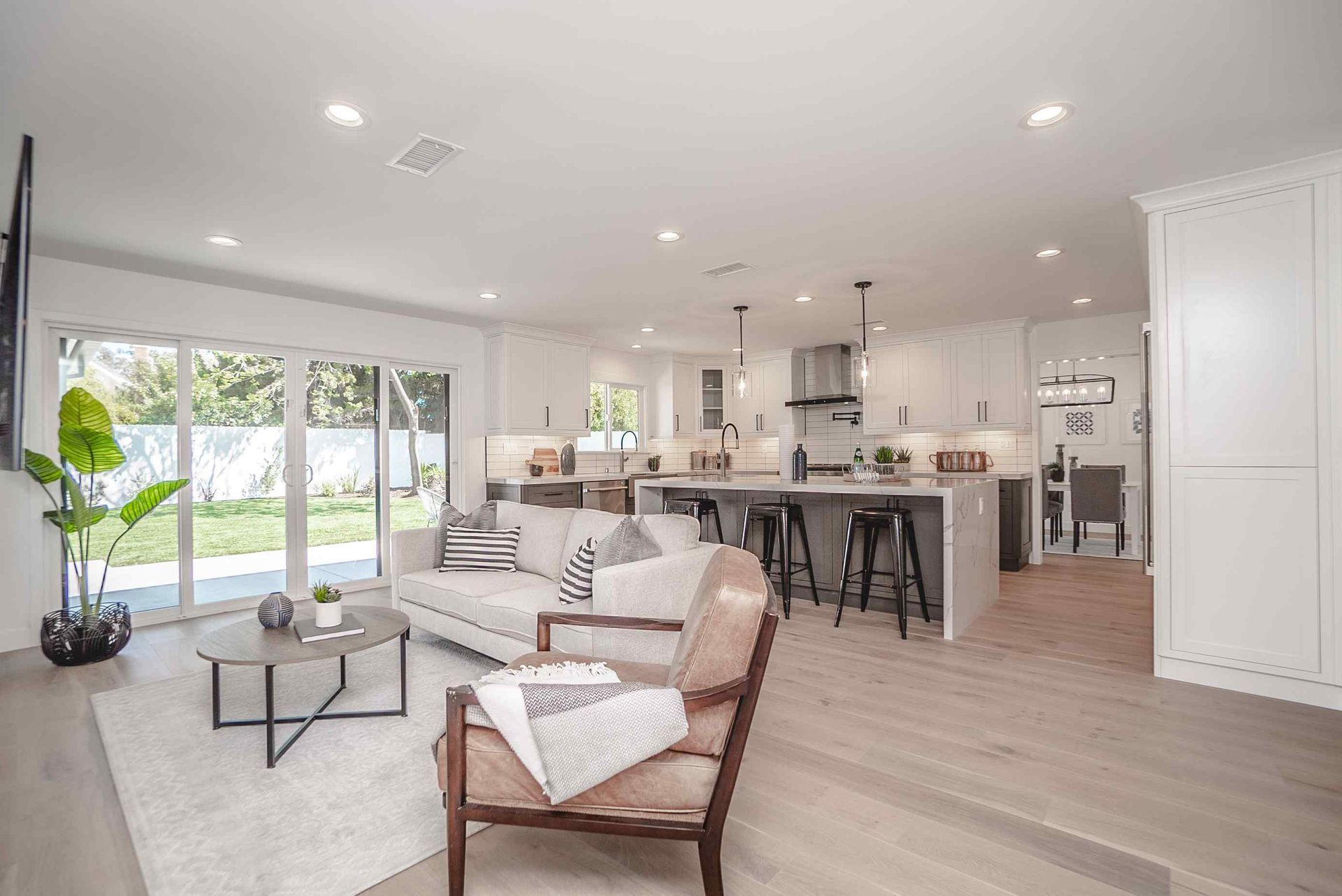 Open-plan living space: white kitchen, grey island with stools, sofa, armchair, sliding glass door to yard.