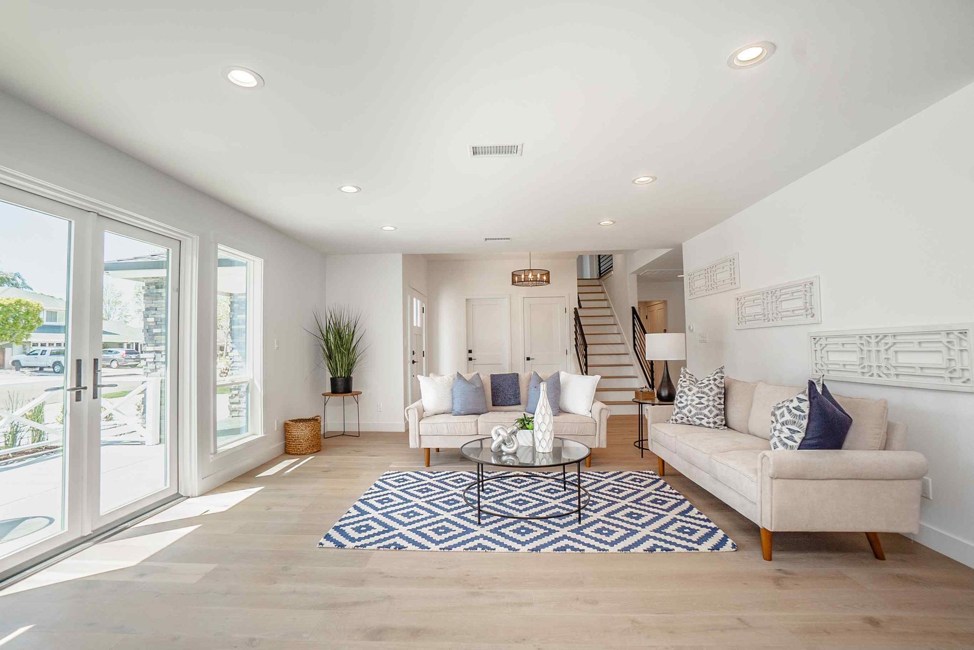 Bright living room with white walls, beige sofas, blue patterned rug, and large windows.