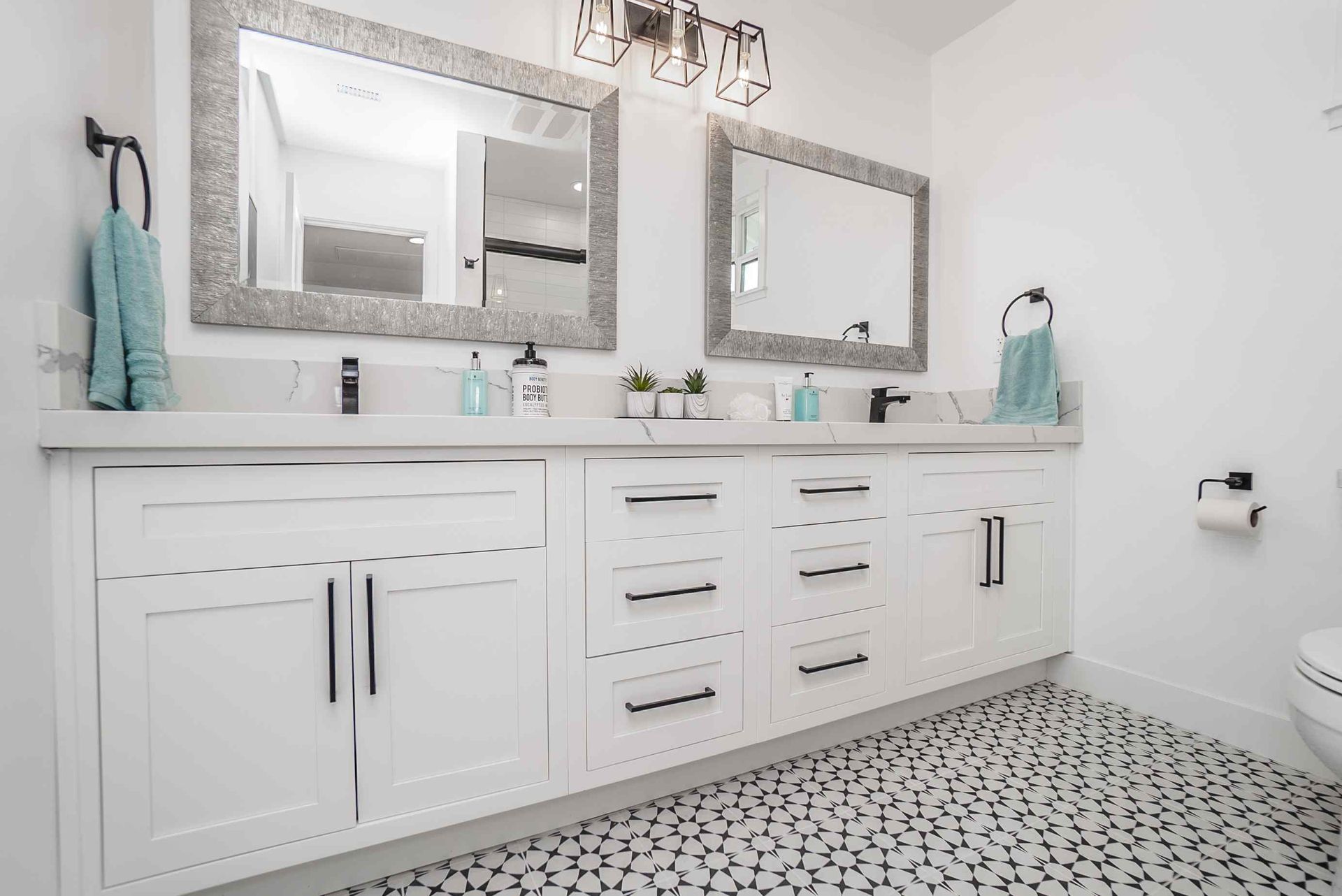 White bathroom with dual sinks, large mirrors, black hardware, and pebble tile flooring.
