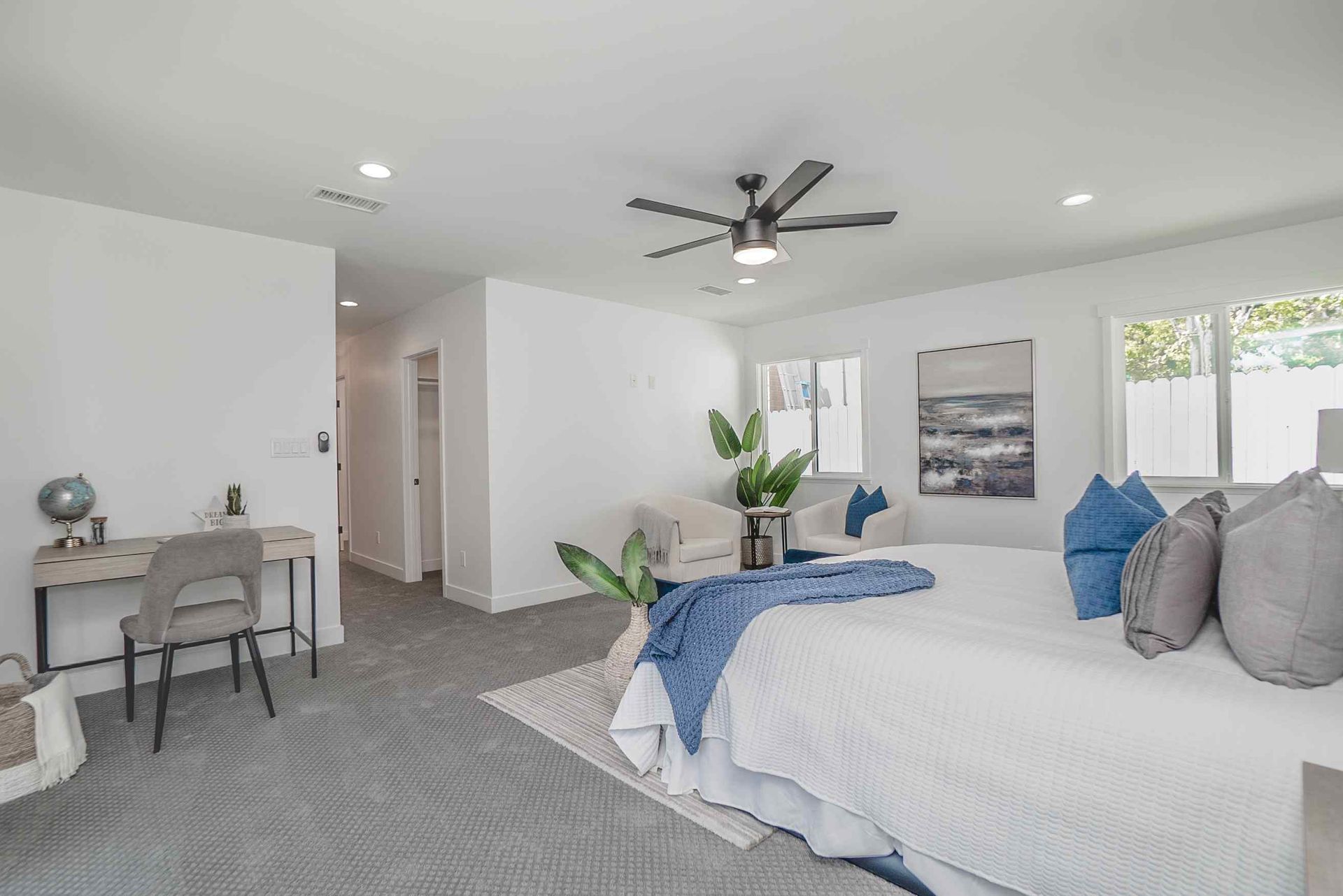 Spacious, modern bedroom with bed, armchair, desk, and artwork. Neutral color scheme with blue accents.