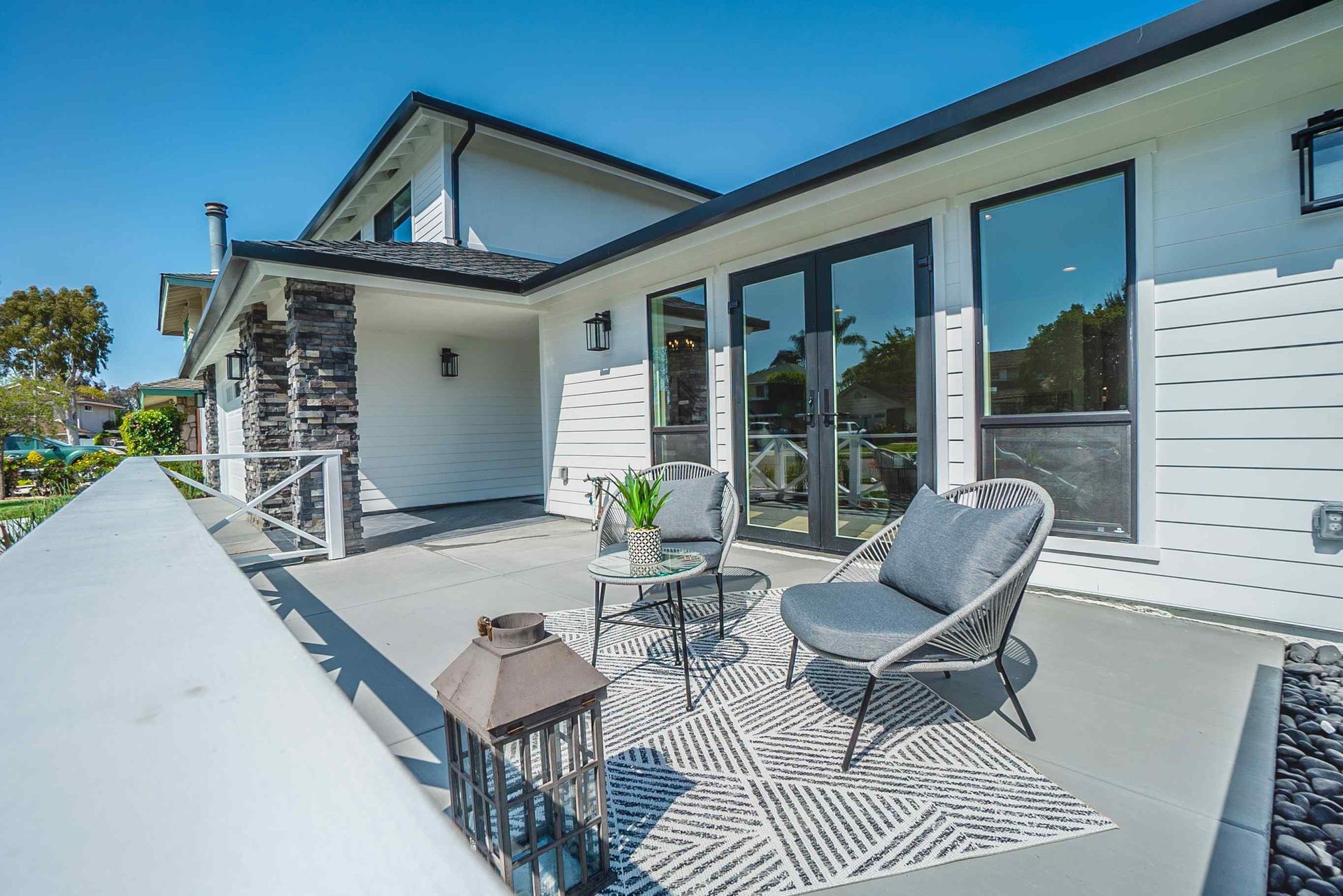 Balcony with gray deck, seating area, and white exterior wall with dark windows and black stone pillar.
