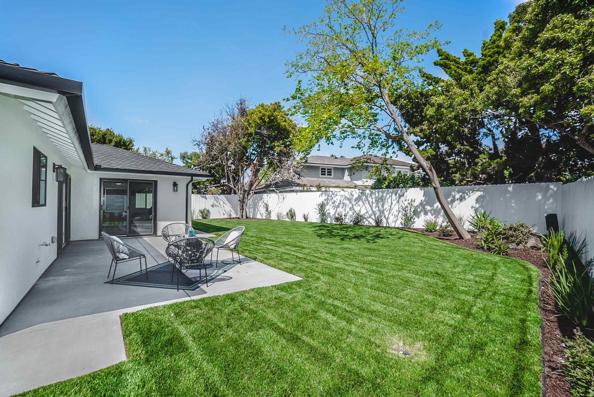 Backyard with green grass, patio, seating, and white walls under a blue sky.
