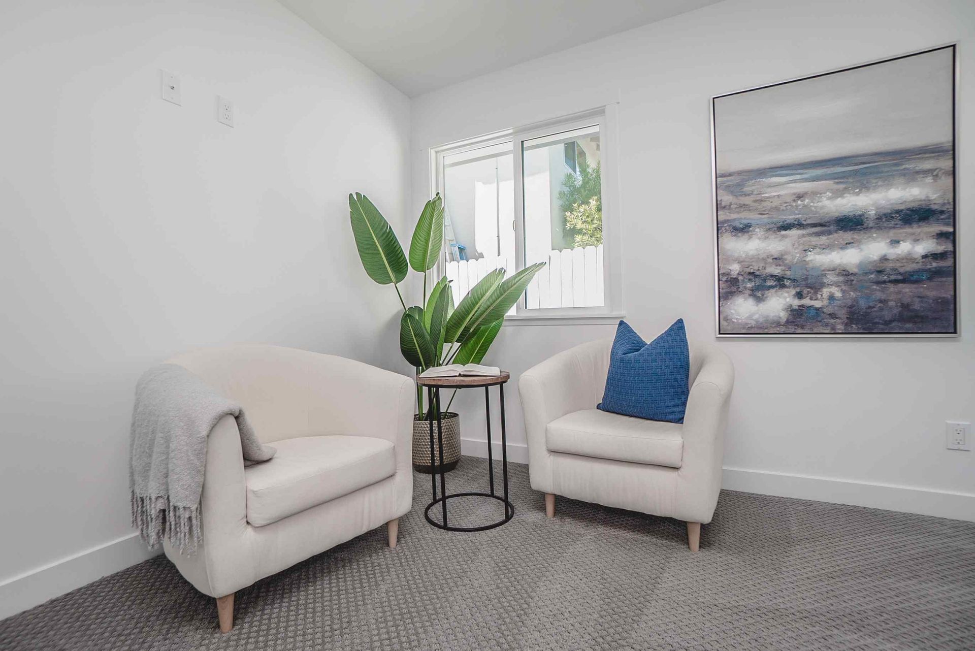 Cozy sitting area with two cream armchairs, a small table, and a large painting on a white wall.