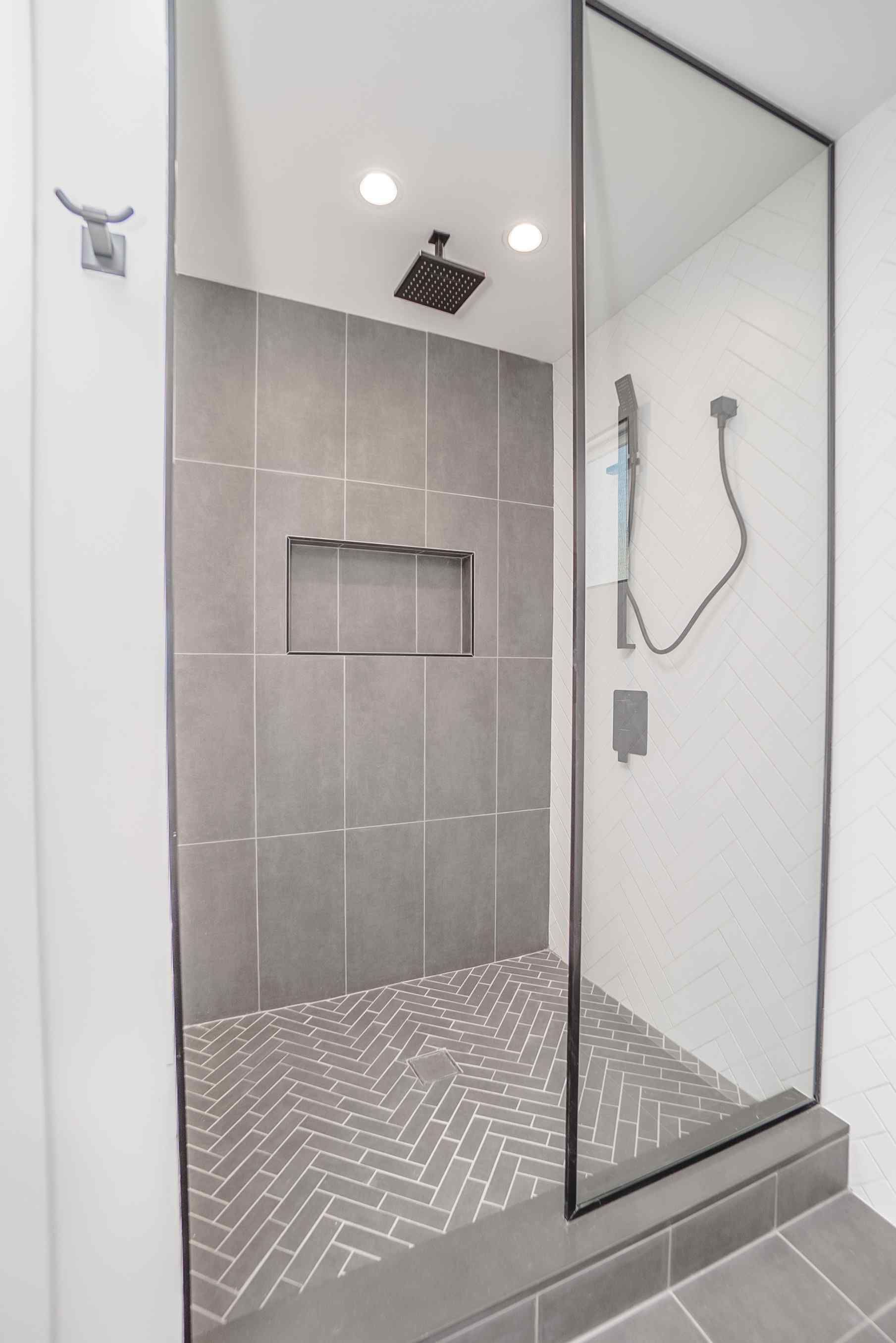 Modern gray and white bathroom with a glass-walled shower, herringbone floor tiles, and a recessed shelf.
