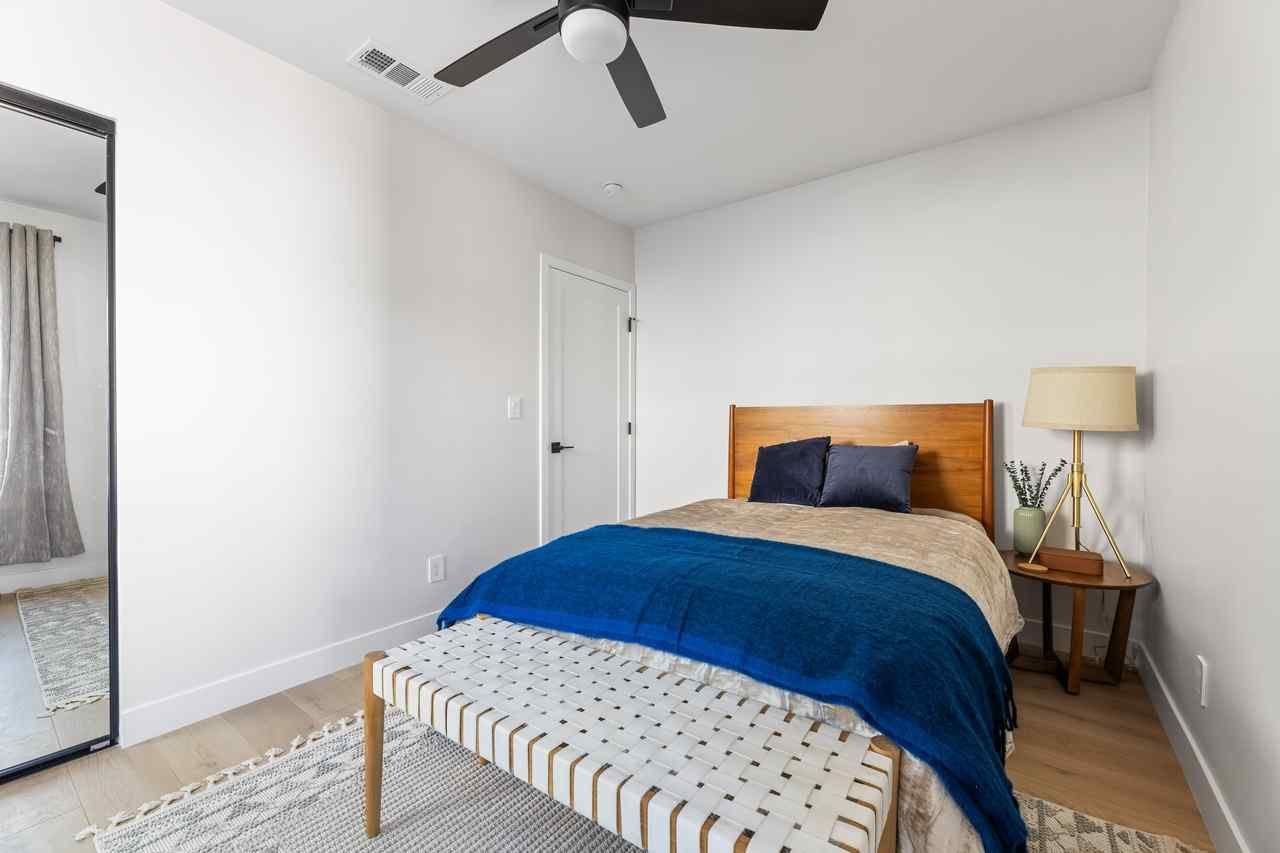 Bedroom with a bed, bench, mirror, and a ceiling fan. Features neutral colors, wood accents, and blue blanket.