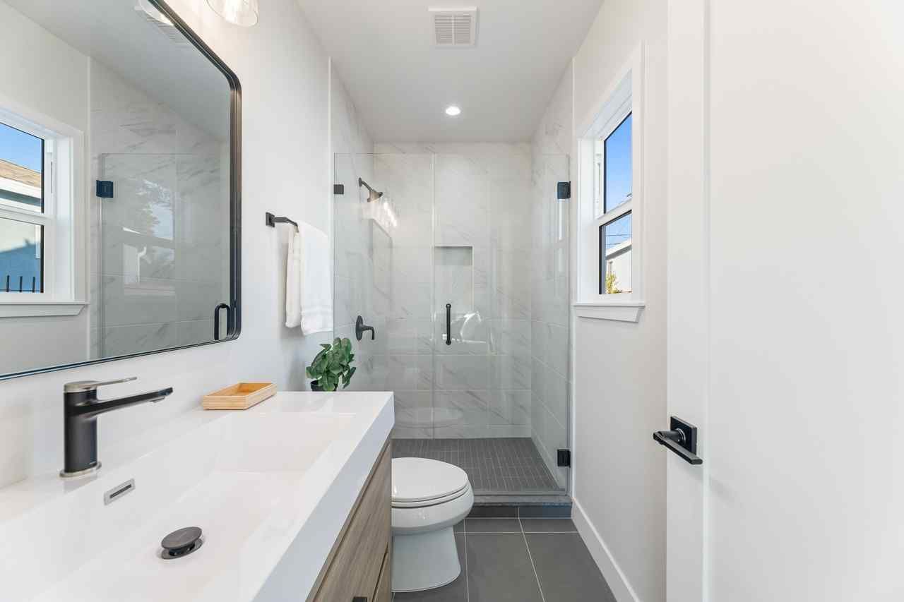 Modern white bathroom with glass shower, toilet, and sink. Black fixtures, gray floor.
