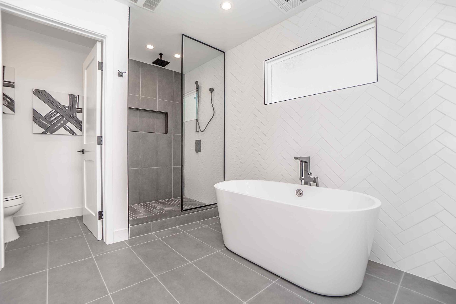 Modern bathroom: white tub, gray shower, white and gray tile, toilet, artwork, and open door.
