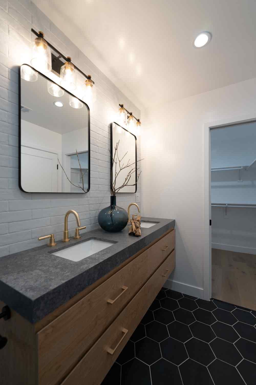 Modern bathroom with double vanity, gold fixtures, black hexagon floor tiles, and white brick wall.