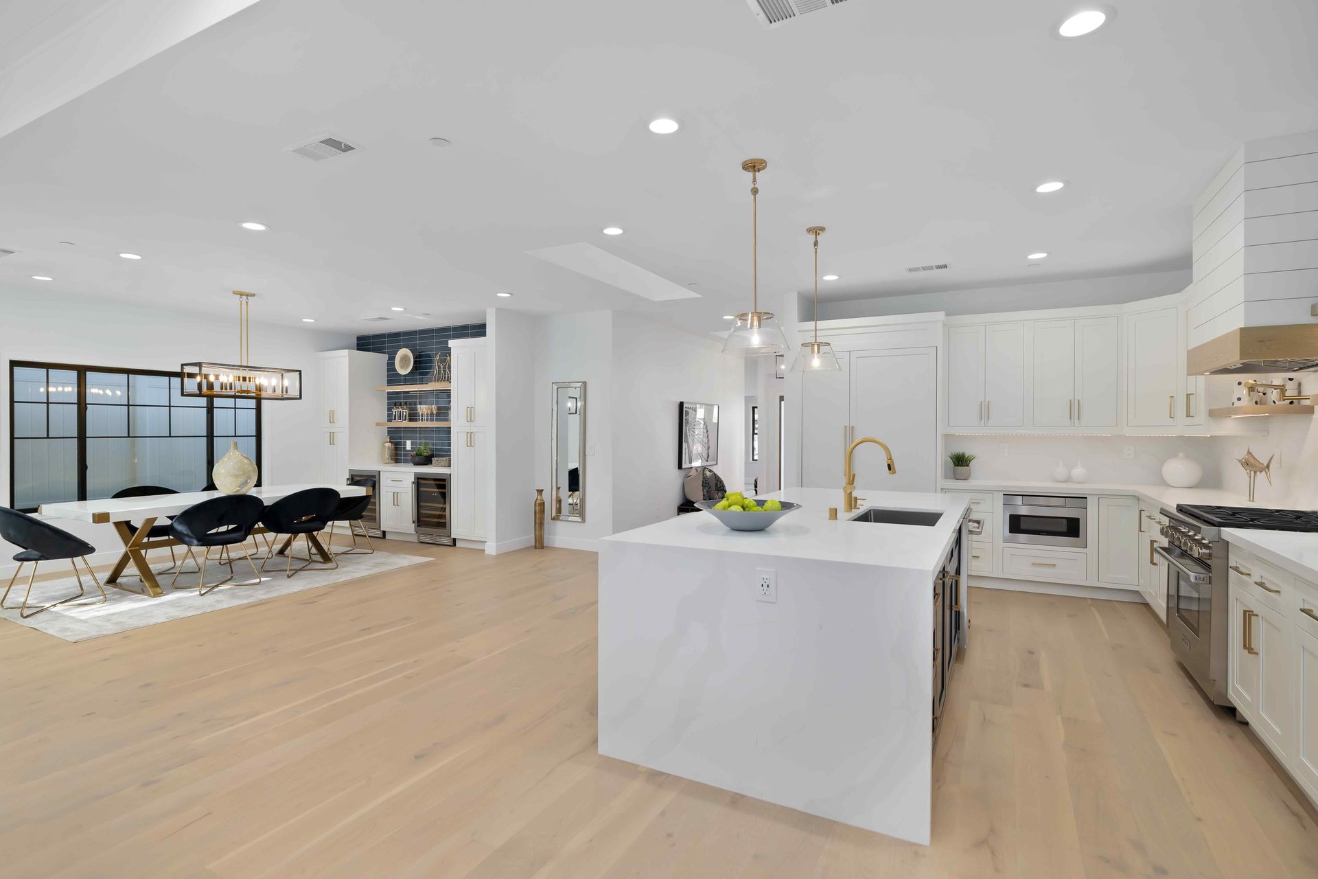 Open-concept kitchen with white cabinets, island, and light wood floors, leading to dining area with black chairs.