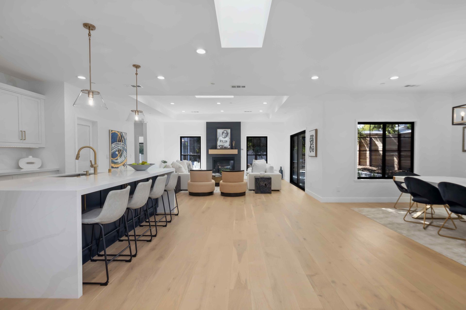 Open-concept modern living space with kitchen, dining, and living room. White walls, light wood floors, and dark accents.