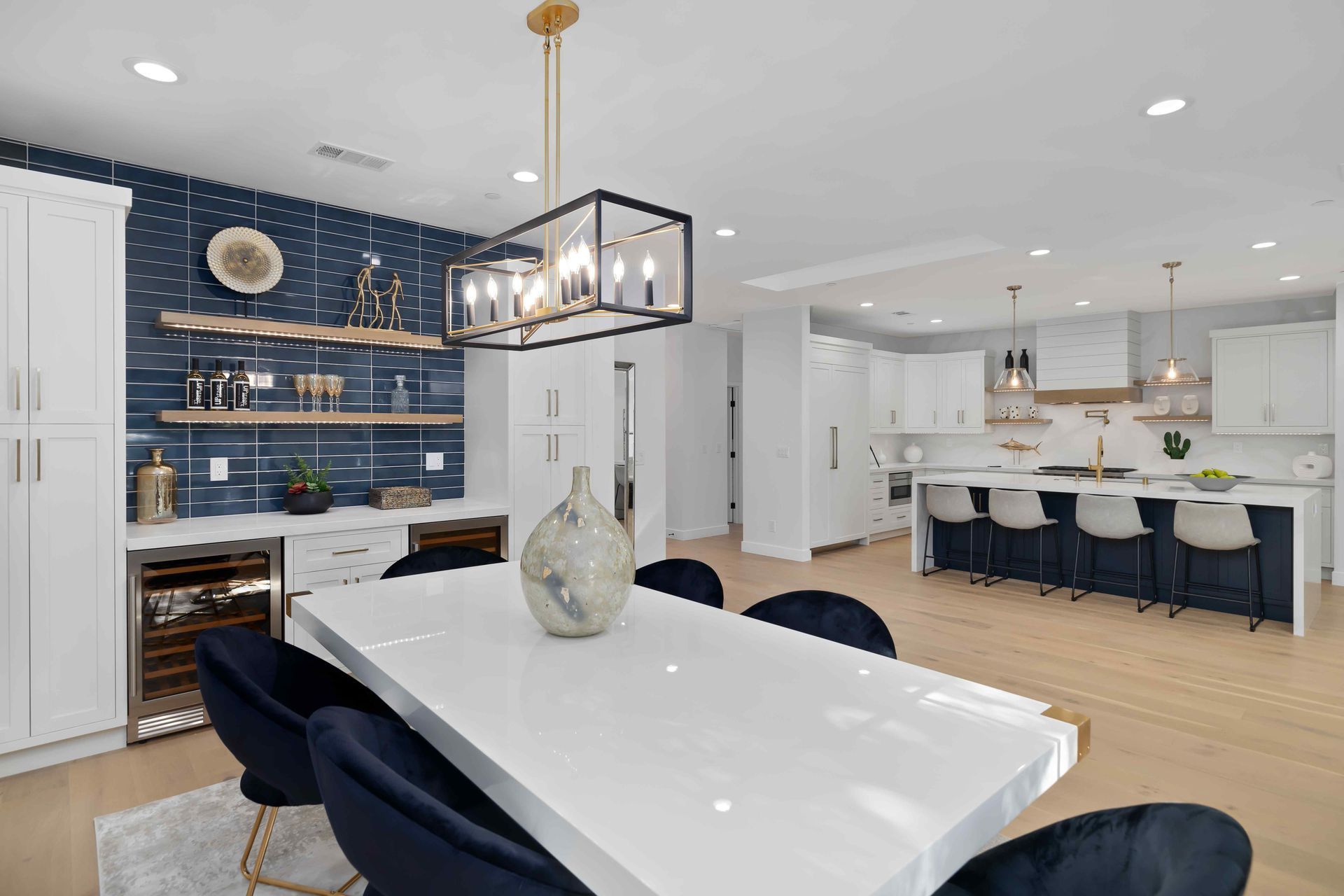 Modern dining room with blue accents: white table, dark blue chairs, open kitchen, and chandelier.