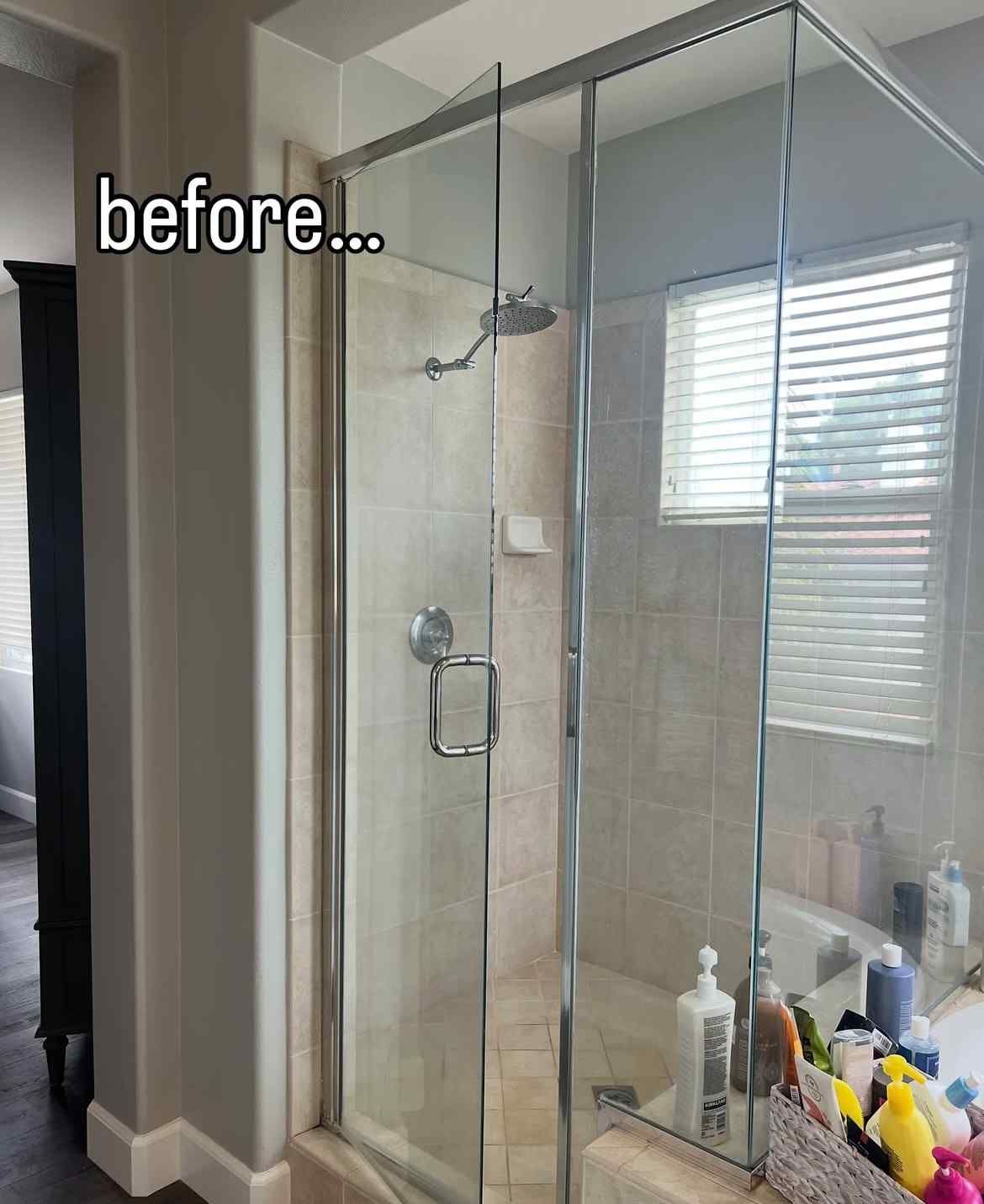 Shower with glass doors and chrome framing in a bathroom setting. Beige tiles and window. 