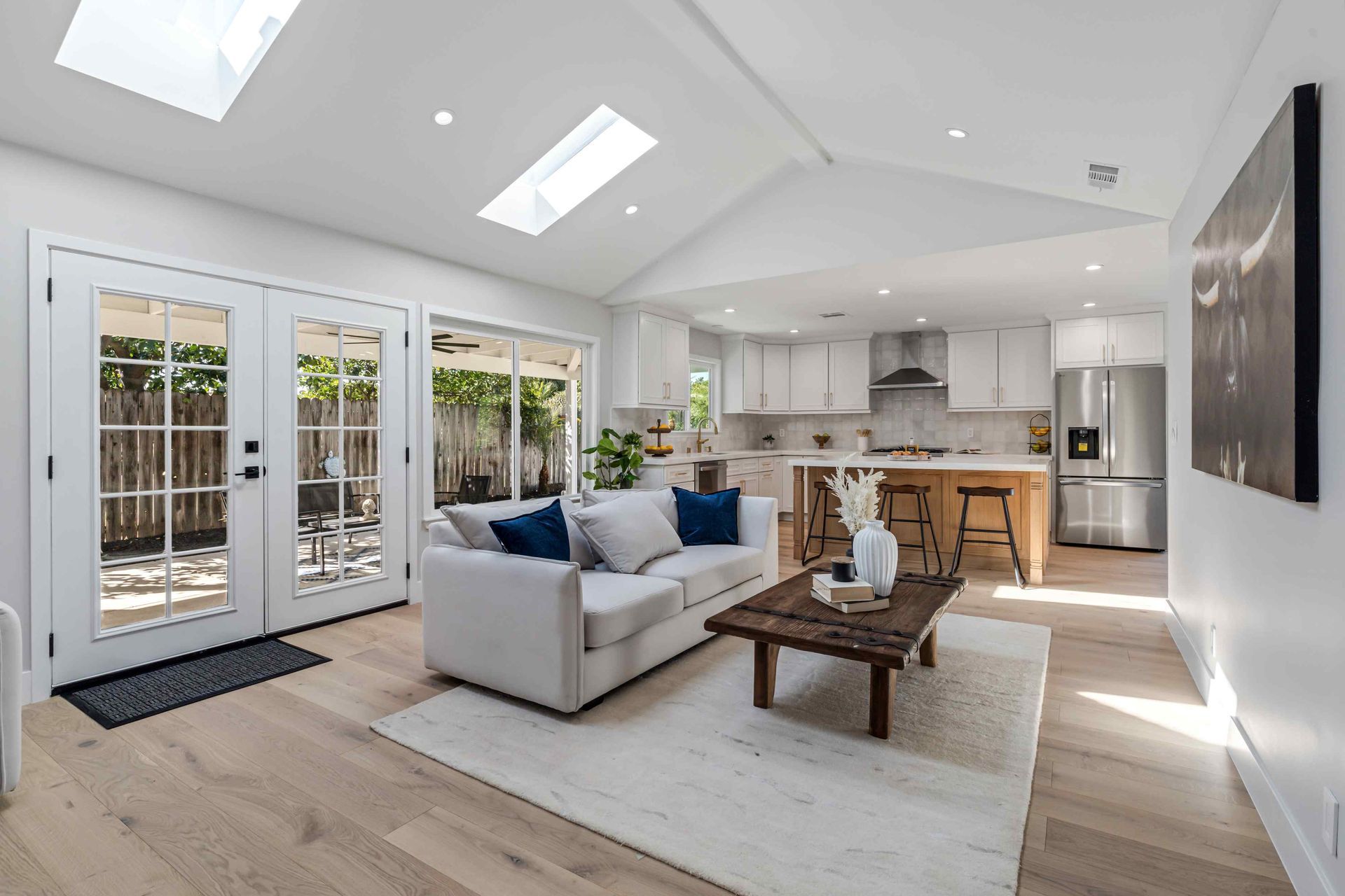Bright open-concept living space with kitchen, white sofa, natural wood floors, and french doors leading to a patio.