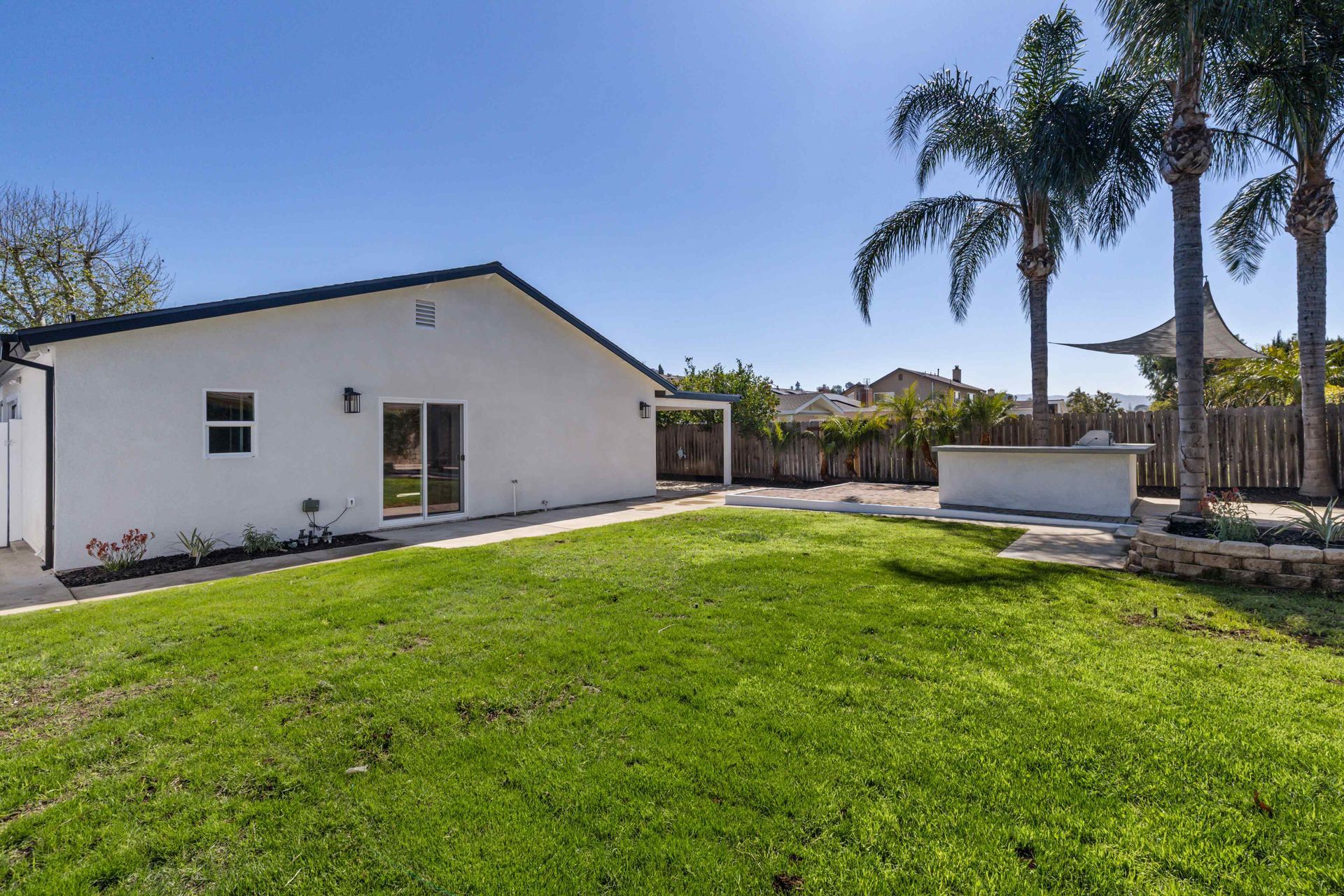 Backyard with white house, green lawn, palm trees, and sunny blue sky.