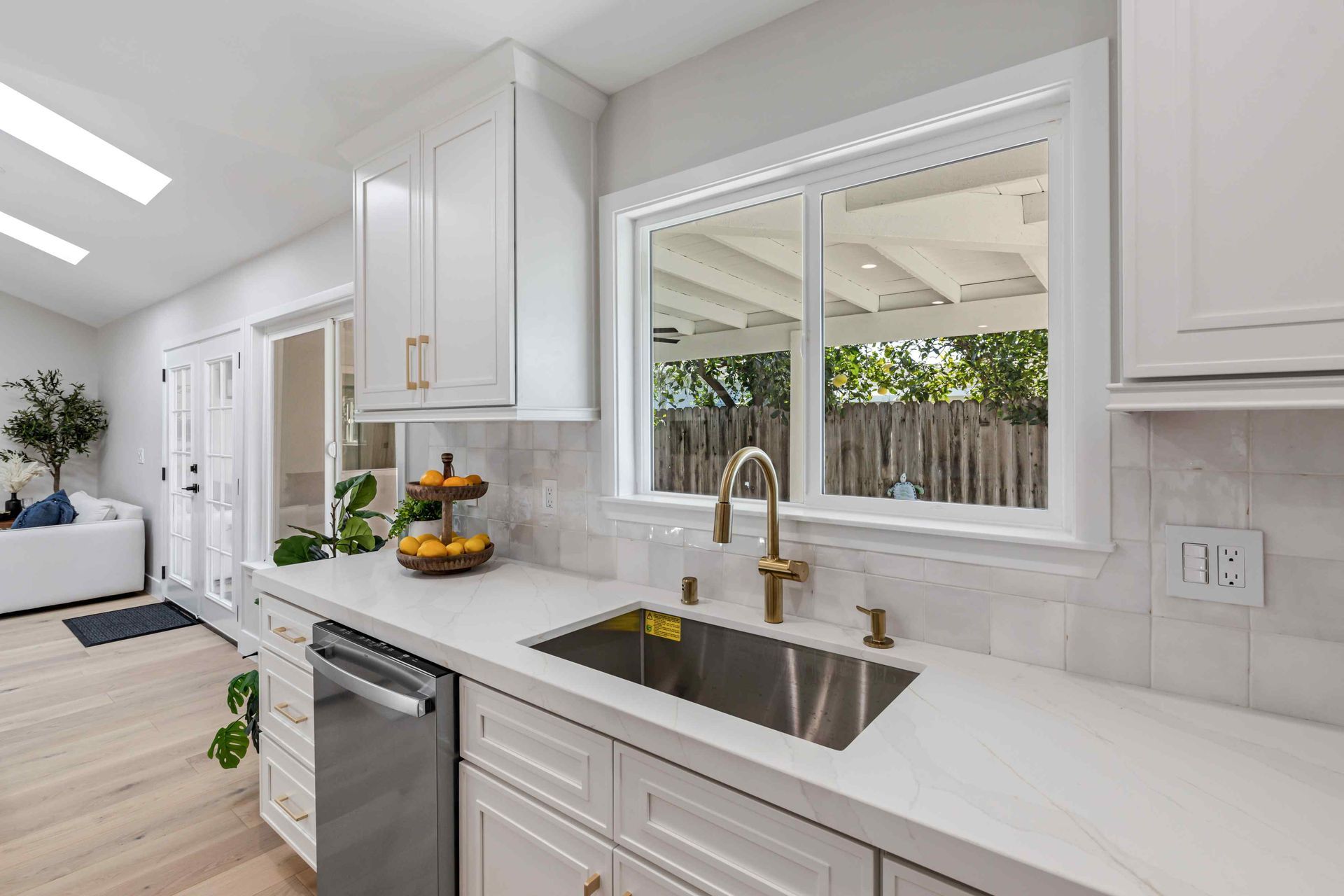 White kitchen with stainless steel sink, gold faucet, and window overlooking backyard.