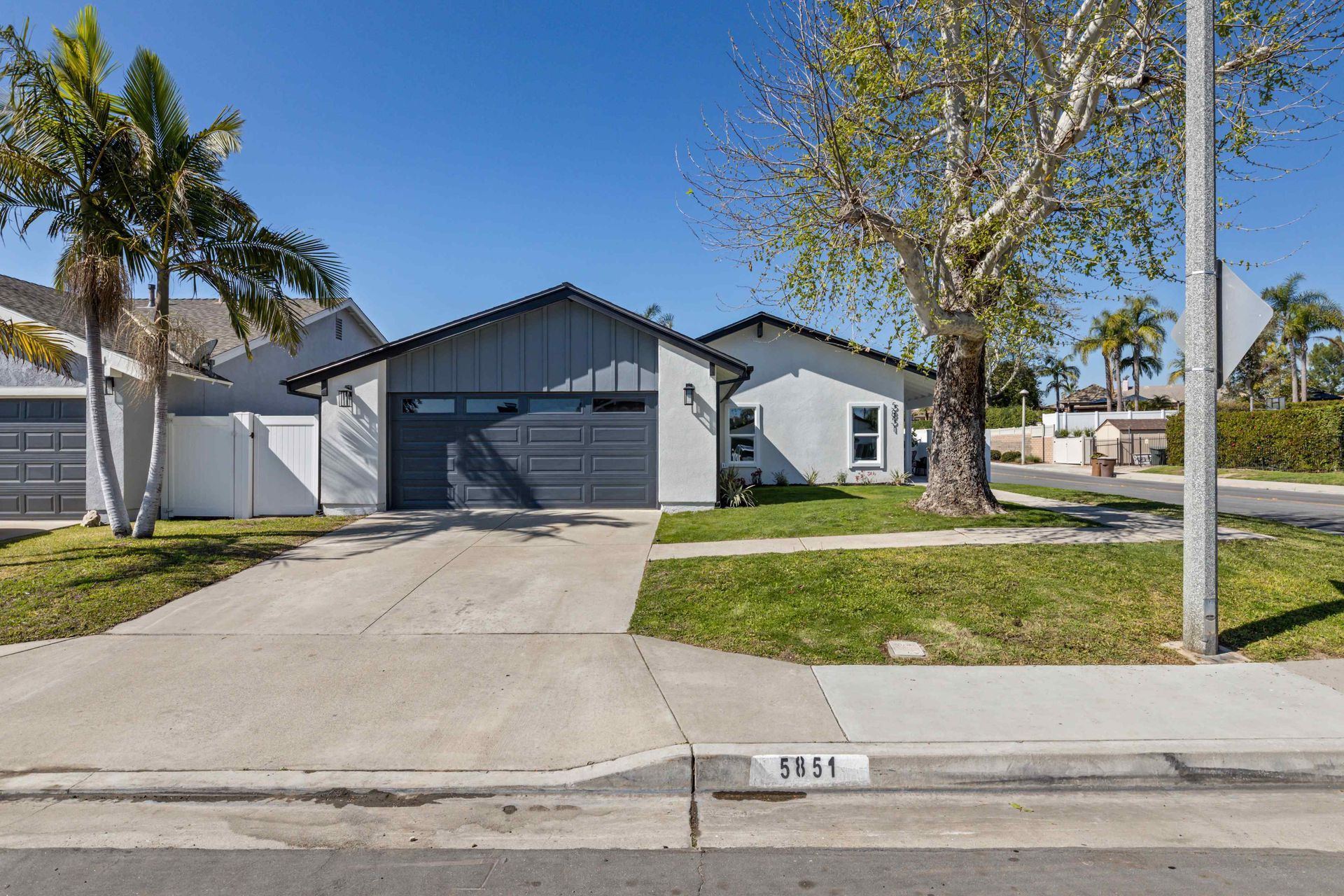 House with grey exterior, driveway, lawn, palm trees, and street sign on a sunny day.