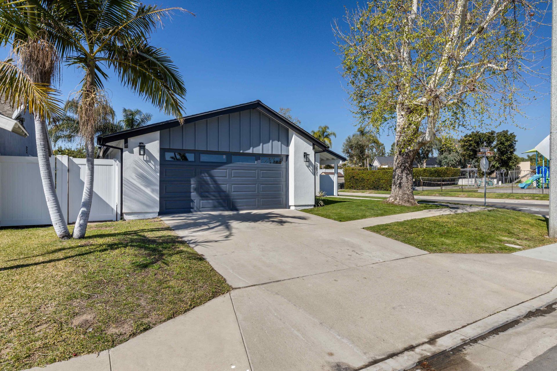 A modern gray house with a driveway, garage door, palm tree, and grass lawn under a blue sky.