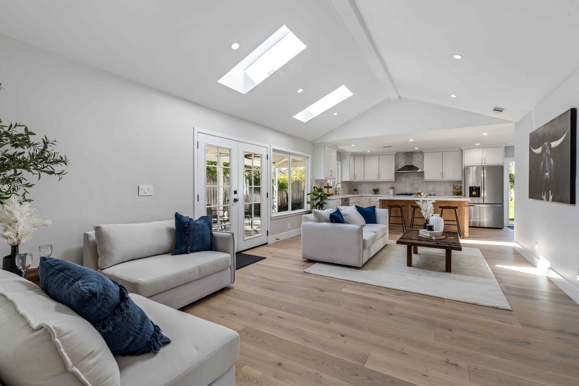 Bright, open living room with skylights, sofas, and a kitchen. Light wood floors and white walls.
