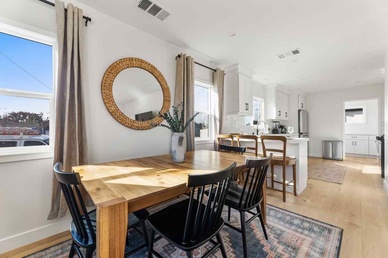 Dining room with wooden table, black chairs, wicker mirror, and open kitchen with light wood floors.