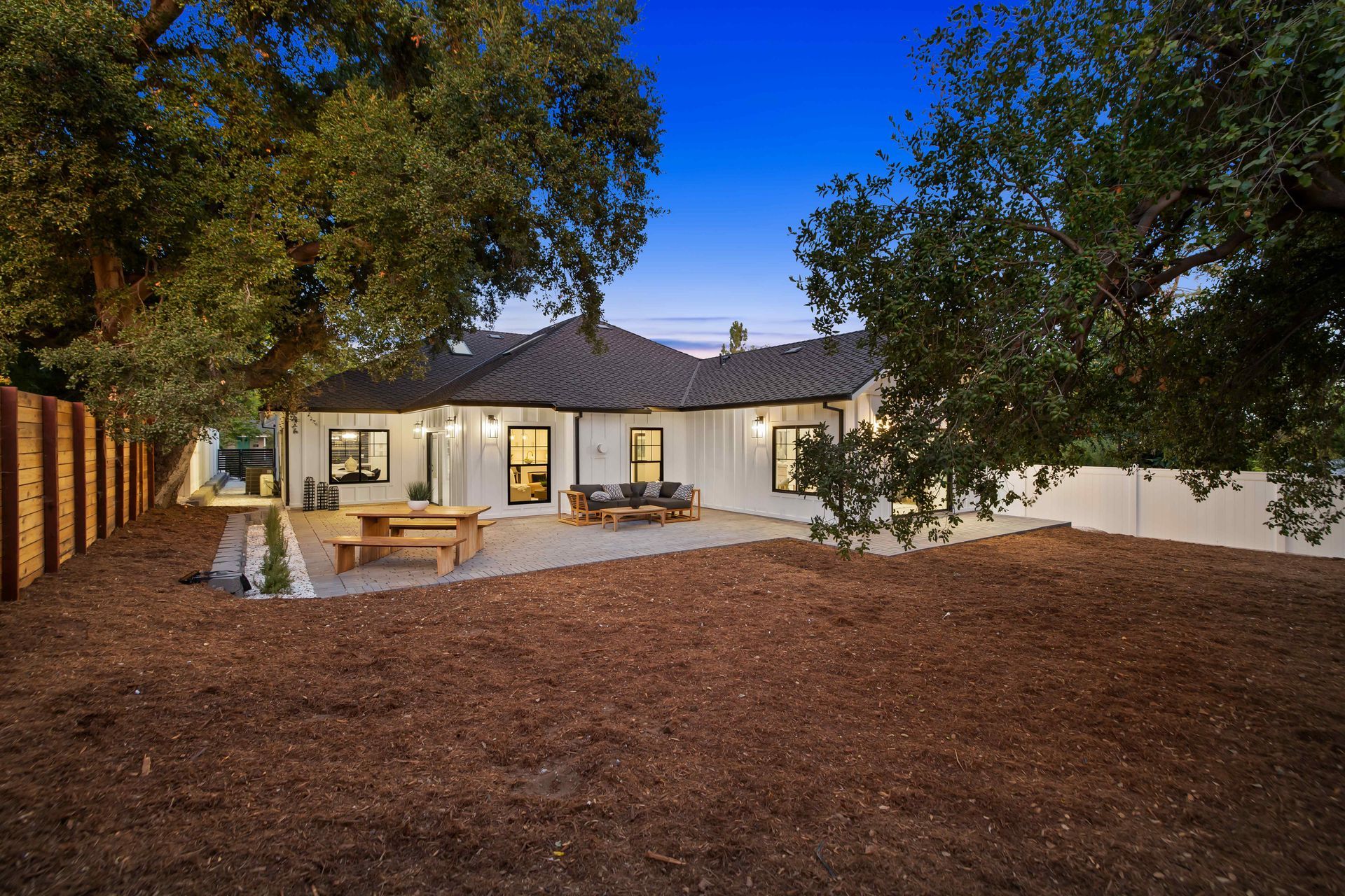 White house with black trim, wooden fence, backyard with mulch, picnic table, and trees.