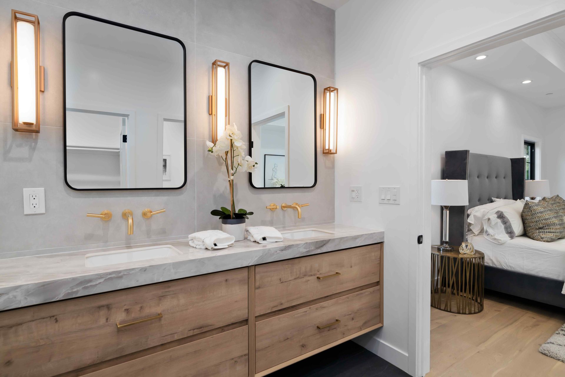 A modern bathroom with double sinks, mirrors, and a wooden vanity. Bedroom visible in the doorway.
