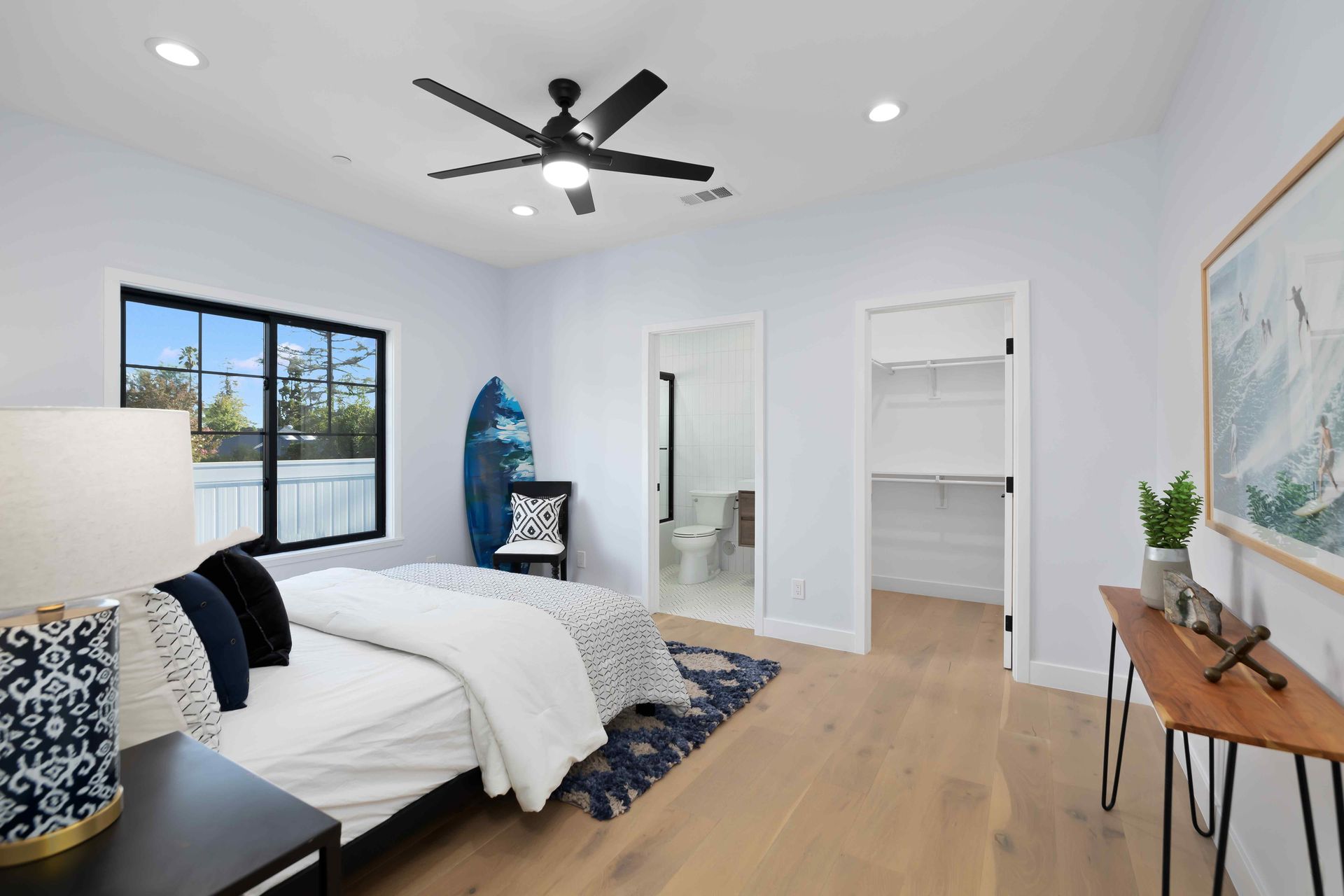 Bedroom with bed, surfboard, and bathroom entrance. Light wood floors, light blue walls, large window, and ceiling fan.