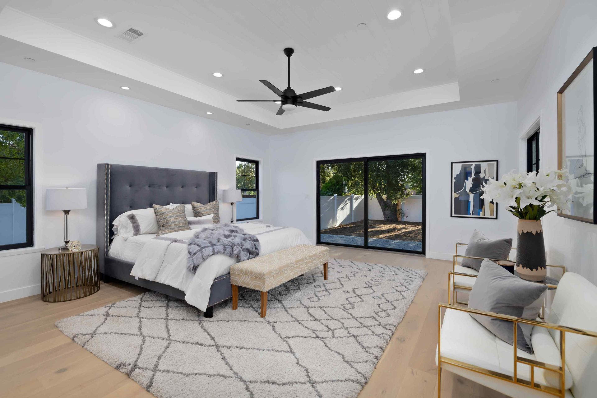 Spacious white bedroom with gray bed, bench, rug, and chairs, plus black windows and door.