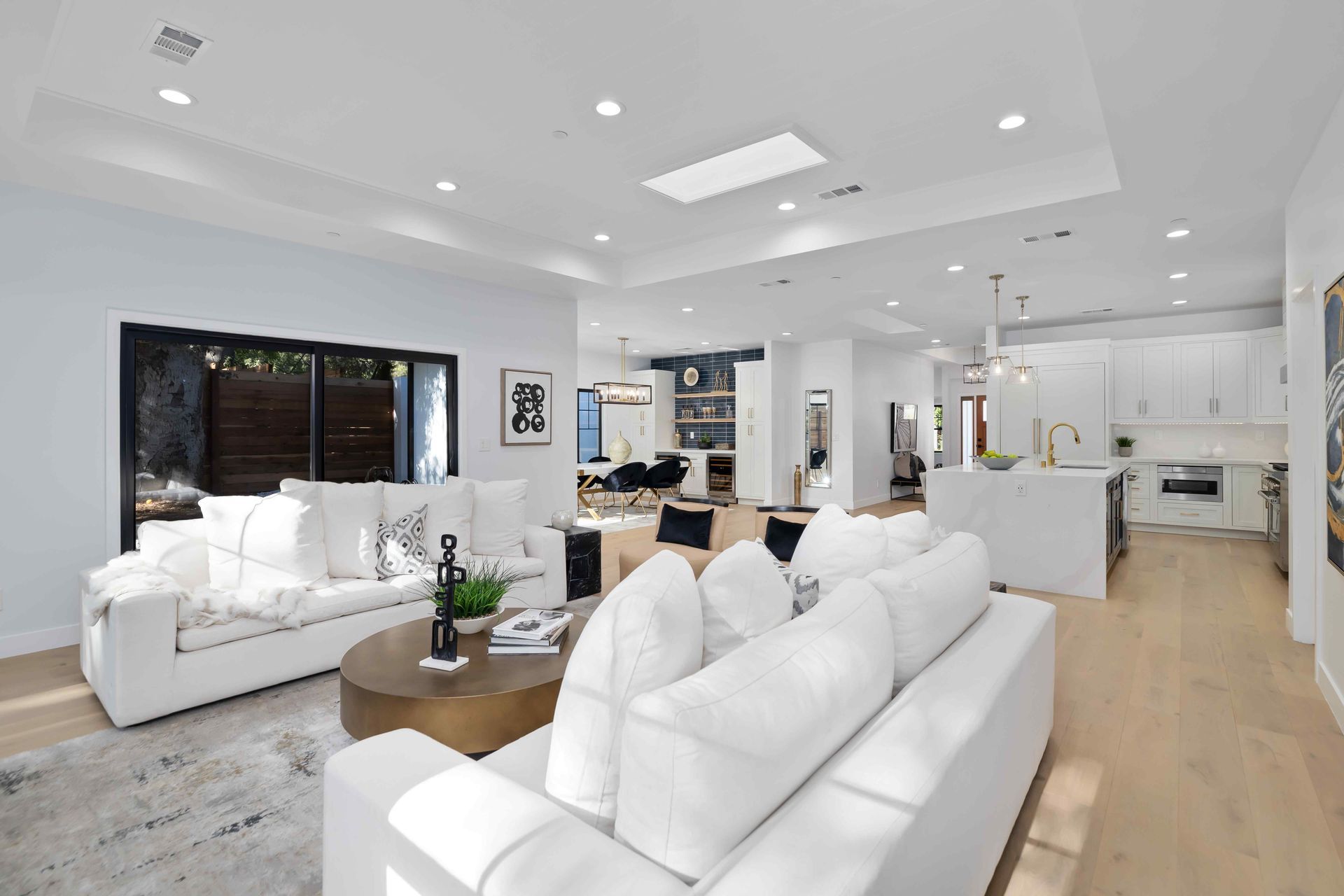 Bright, open-plan living room with white sofas, a round coffee table, and a kitchen visible in the background.